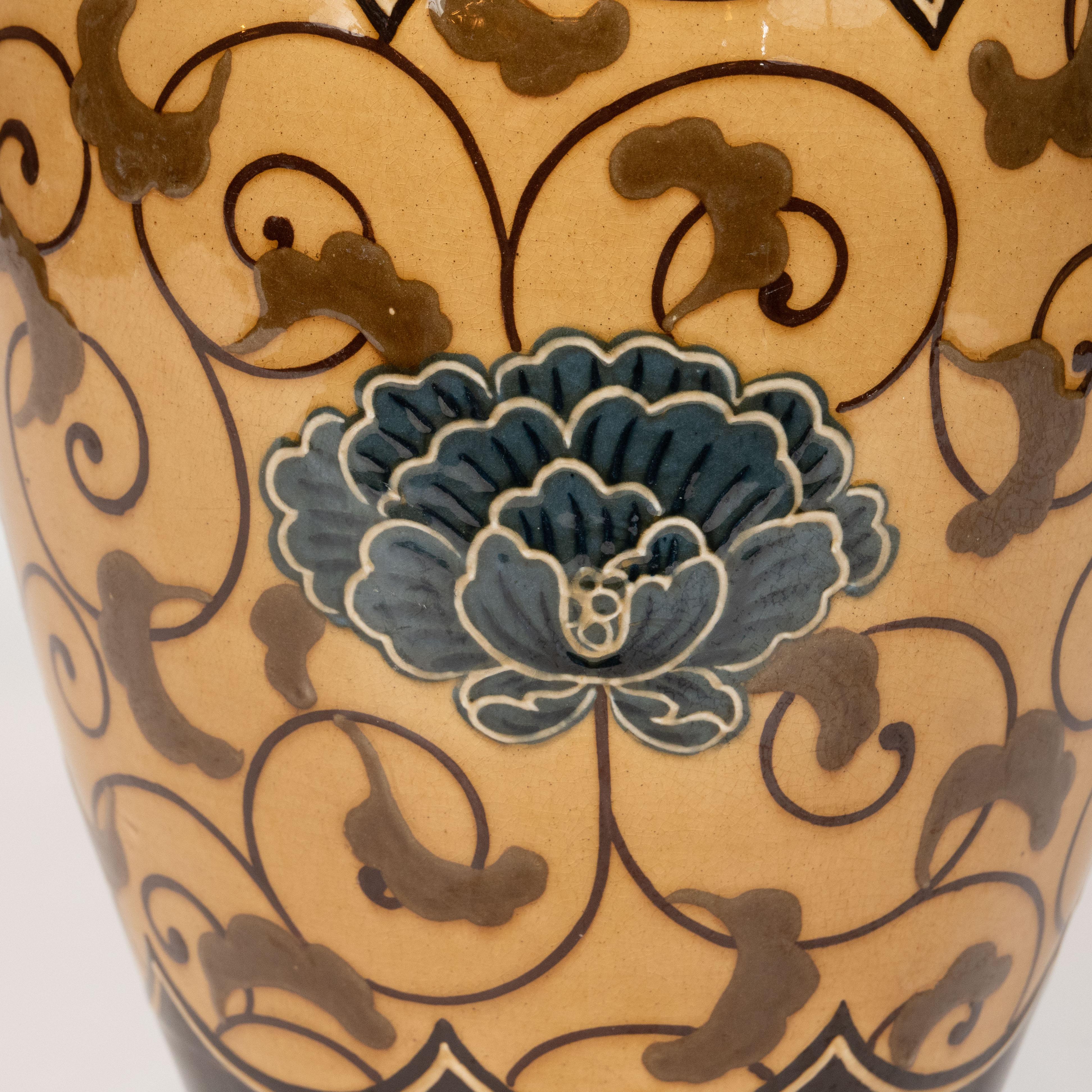 This elegant Art Nouveau vase was realized in the England by the storied maker Wedgwood circa 1890. It features Art Nouveau stylized foliate patterns in caramel and crimson with an indigo hued flower in the center. The ceramic vase tapers slightly