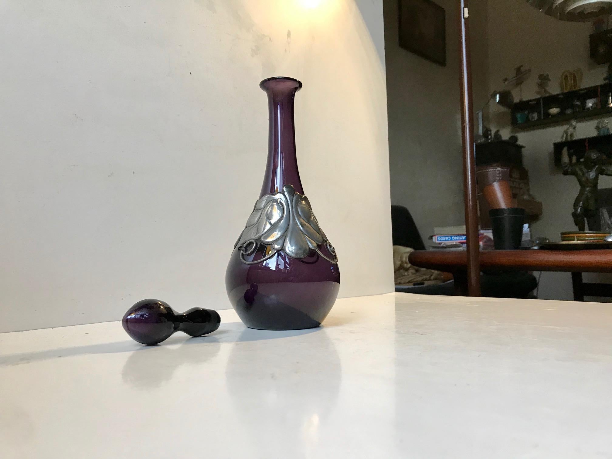 Organically shaped hand blown decanter with a floral decoration in pewter. This decanter was made at Holmegaard in Denmark in 1923. The Jugendstil pewter belt was created by a Danish silversmith in the same period. This item is catalogued and