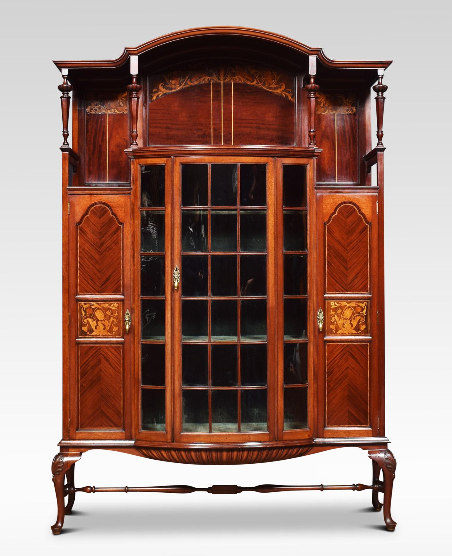 An Art Nouveau inlaid display cabinet probably by Shapland & Petter, The arched superstructure supported on four carved tapering columns to the large central glazed bowed door opening to reveal upholstered shelved interior. Flanked by mahogany
