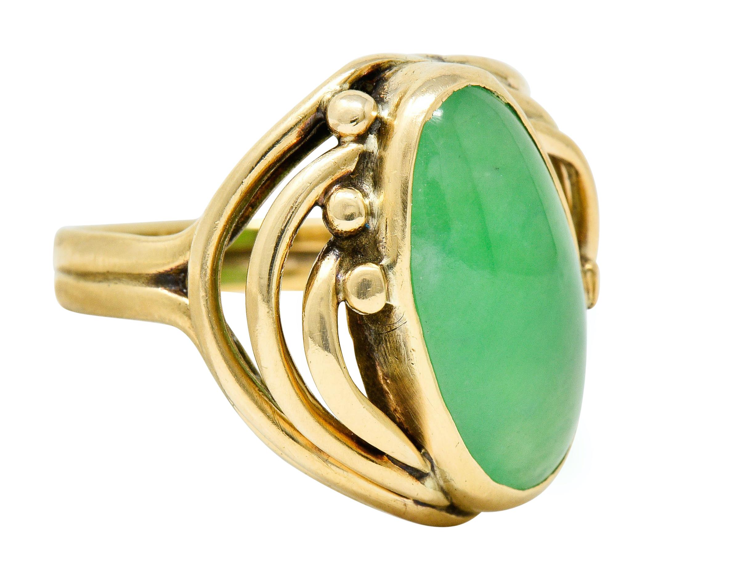 Band ring centers an elongated oval jadeite jade cabochon; measuring approximately 16.5 x 19.2 mm

Translucent with uniform green color with no indications of impregnation; type A jade

Surrounded by arched whiplash branches terminating as gold bead