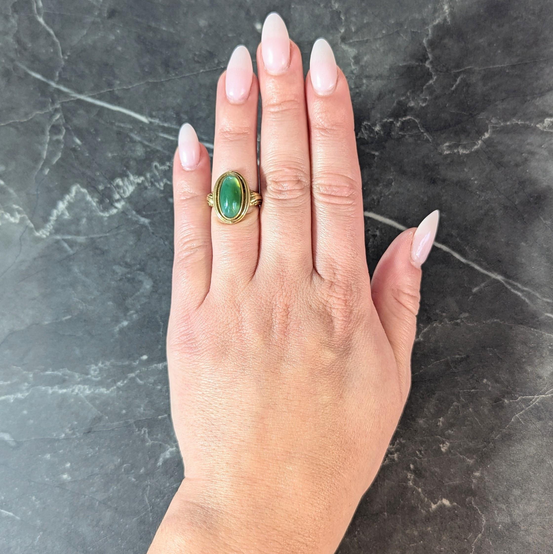 Centering an oval shaped jadeite jade cabochon measuring 7.0 x 14.0 mm
Translucent light to medium green in color and bezel set
With a fluted halo surround accented by gold beading
Flanked with grooved sweeping scroll motif shoulder
Tested as 18
