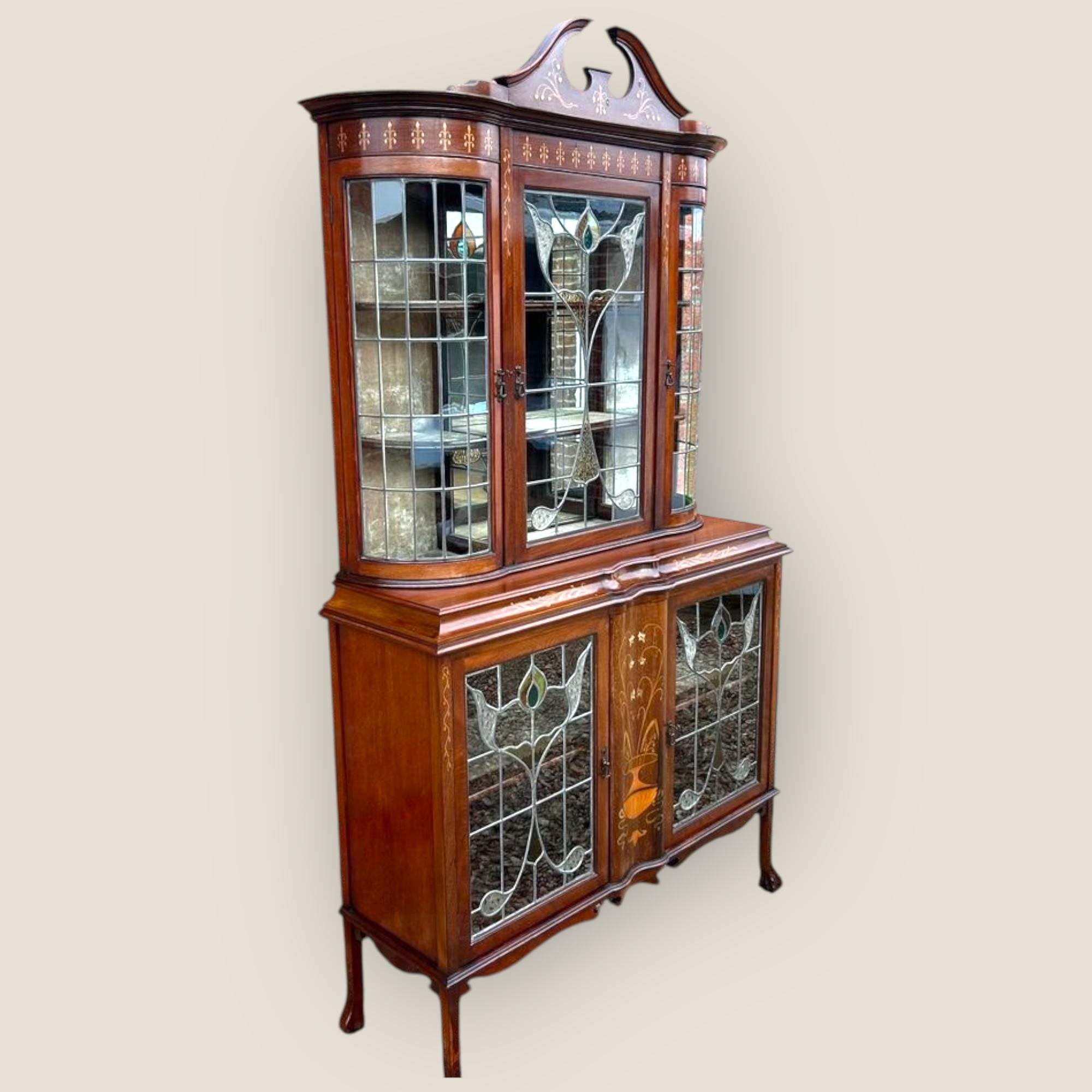 An Edwardian art nouveau mahogany display cabinet. Featuring lead light glazed doors including floral designs and an inlaid broken style pediment to the top. Inlaid from top to bottom with floral patterns of marquetry, copper and brass. The top