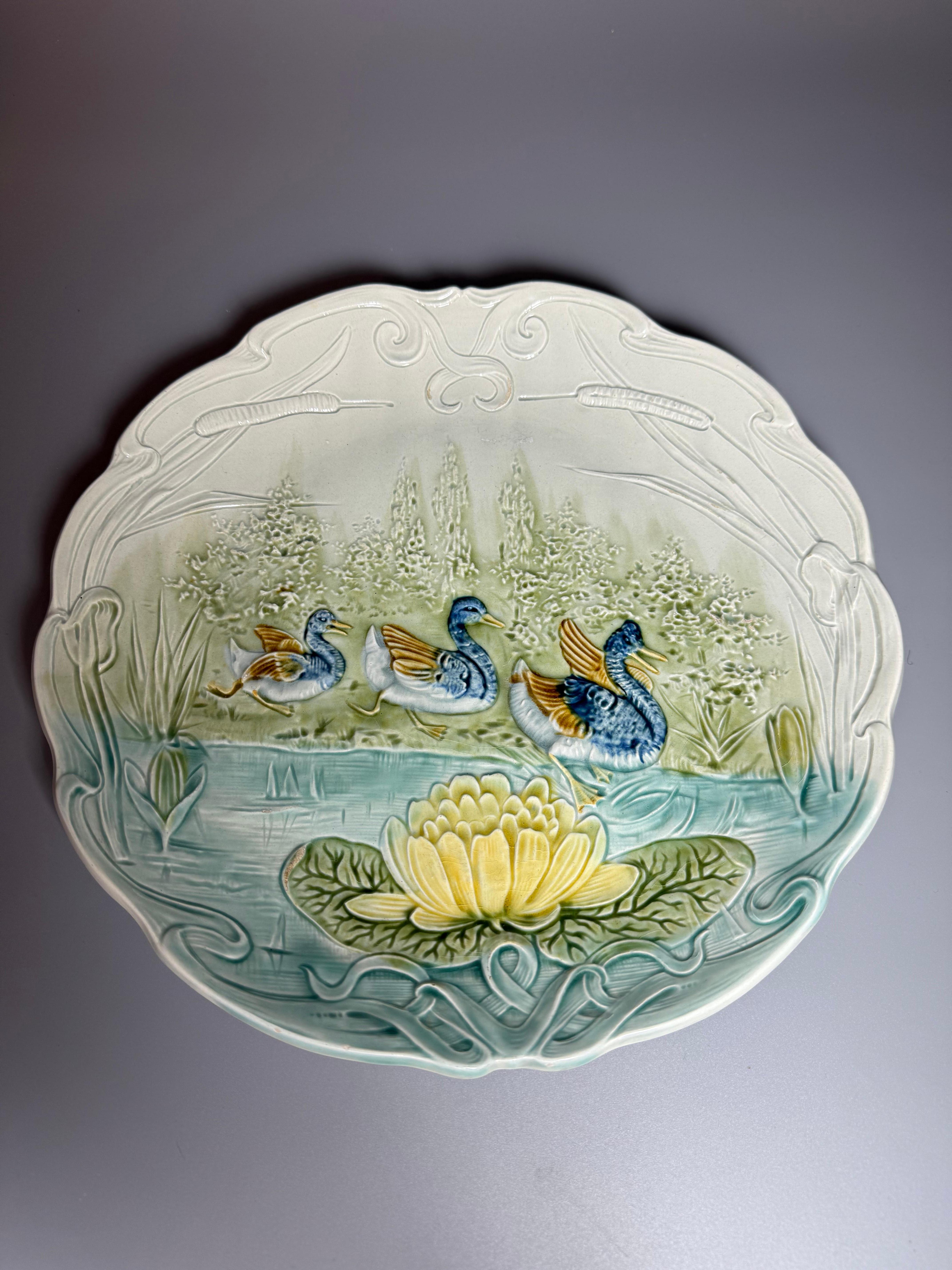 A late 19th to early 20th-century Majolica wall plate featuring a scene with swans on a lake and a water lily in the foreground. 
This specific plate is described as having a charming scalloped edge and embossed details, with a shaped rim featuring