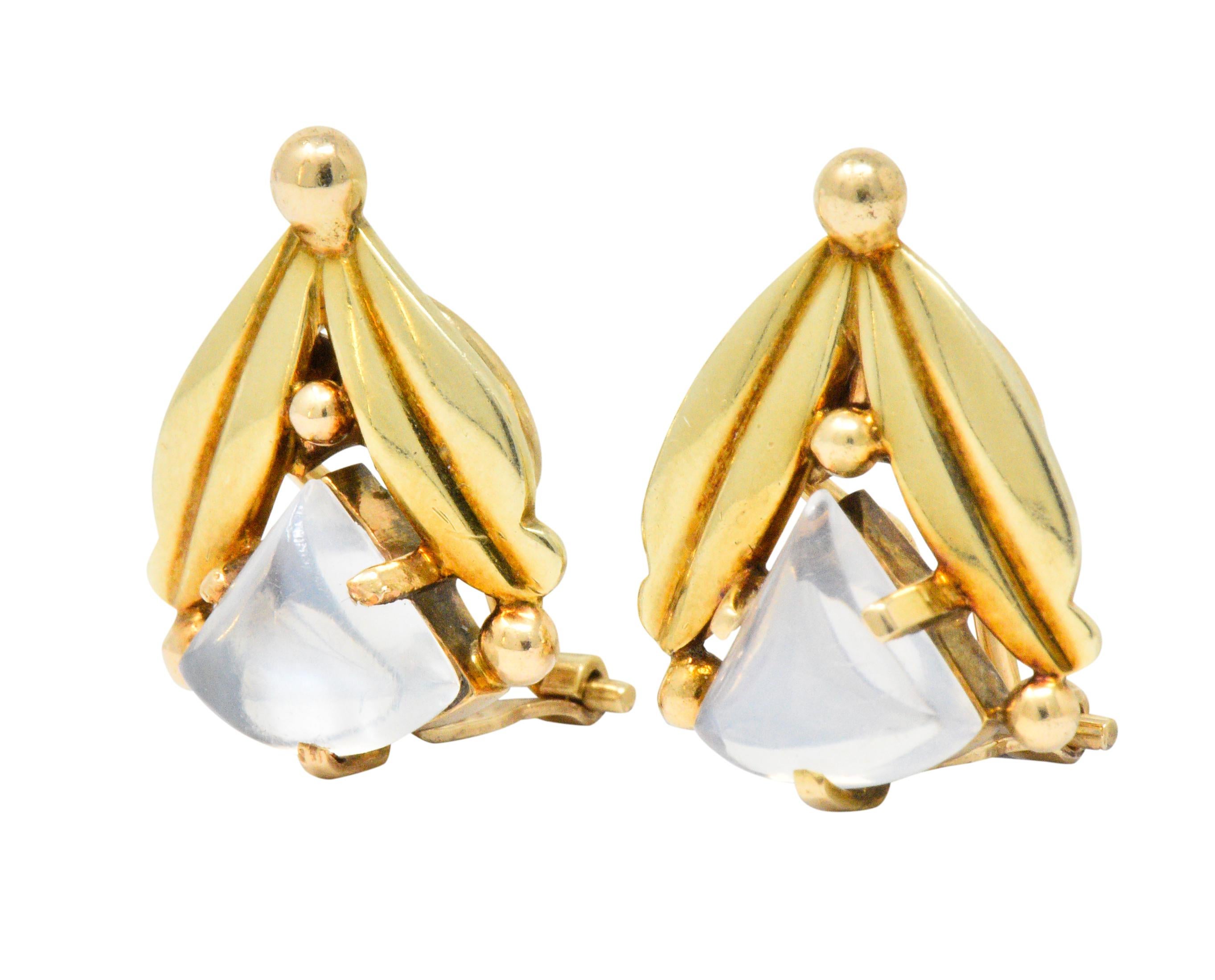 Earrings designed as two polished gold leaves, deeply grooved, in a V formation

Centering triangular cabochons of moonstone, measuring approximately 9.3 x 9.8 mm, translucent with billowy blue adularescence

Accented by polished gold