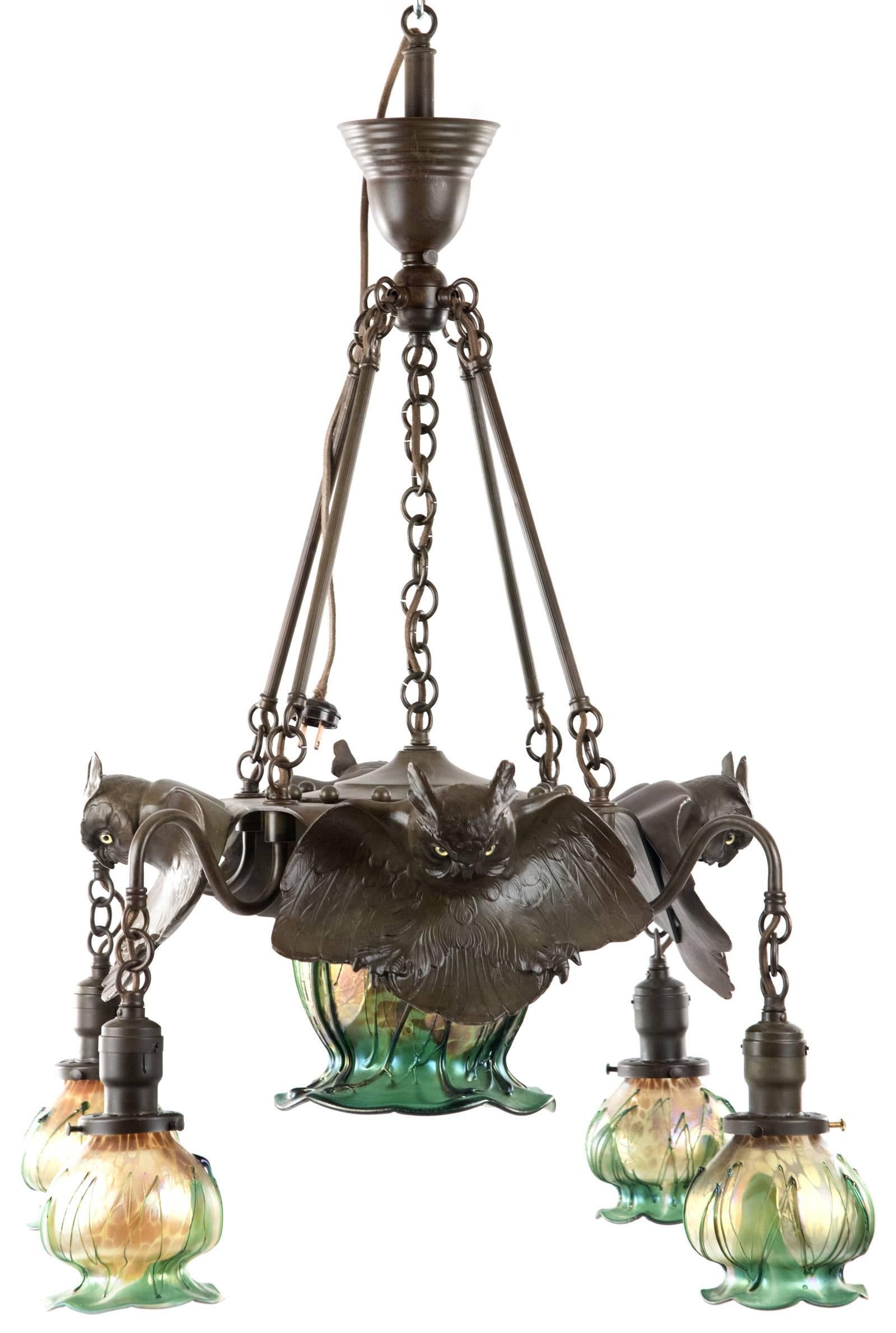 An Art Nouveau style brass chandelier with the hanging support with four mounted and incised owl forms with out-stretched wings and glass eyes, surrounded by four tulip-shaped blown glass shades supported on scrolled arms, above a large central