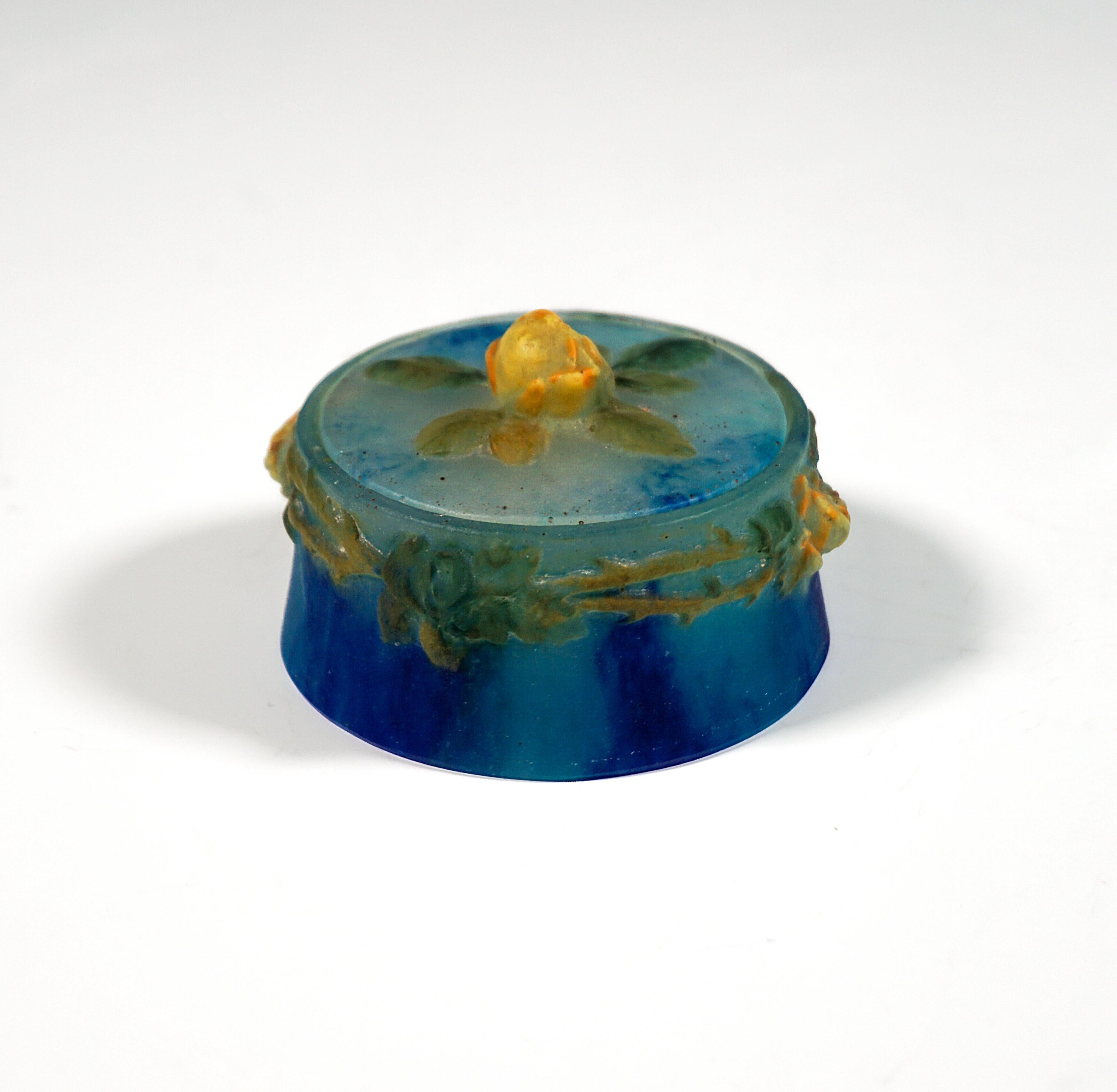 Delicate round lidded box made of translucent glass paste with green, blue and turquoise color inclusions, with slightly conical wall, recessed 
flat lid with yellow rosebud and green leaves as knob, on the upper rim of the container paired