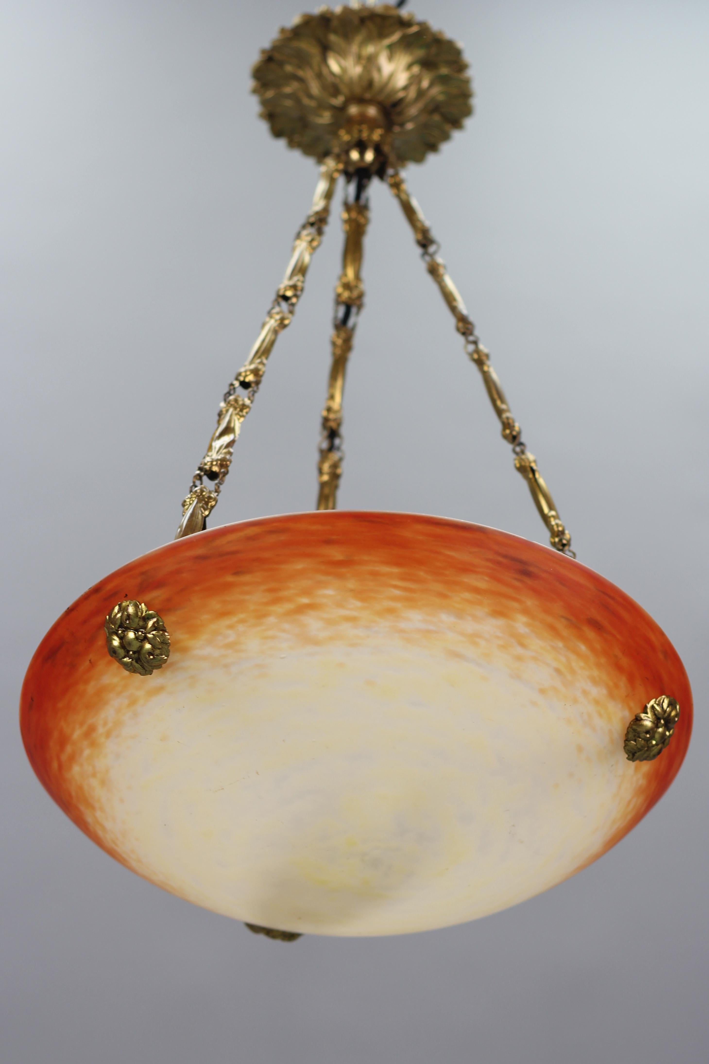 This adorable French Art Nouveau period pendant chandelier features a mottled “Pâte de Verre” art glass shade in orange, yellow, and cream color, signed ”Schneider” by Charles Schneider, hung at an ornate brass and bronze fixture with one socket for