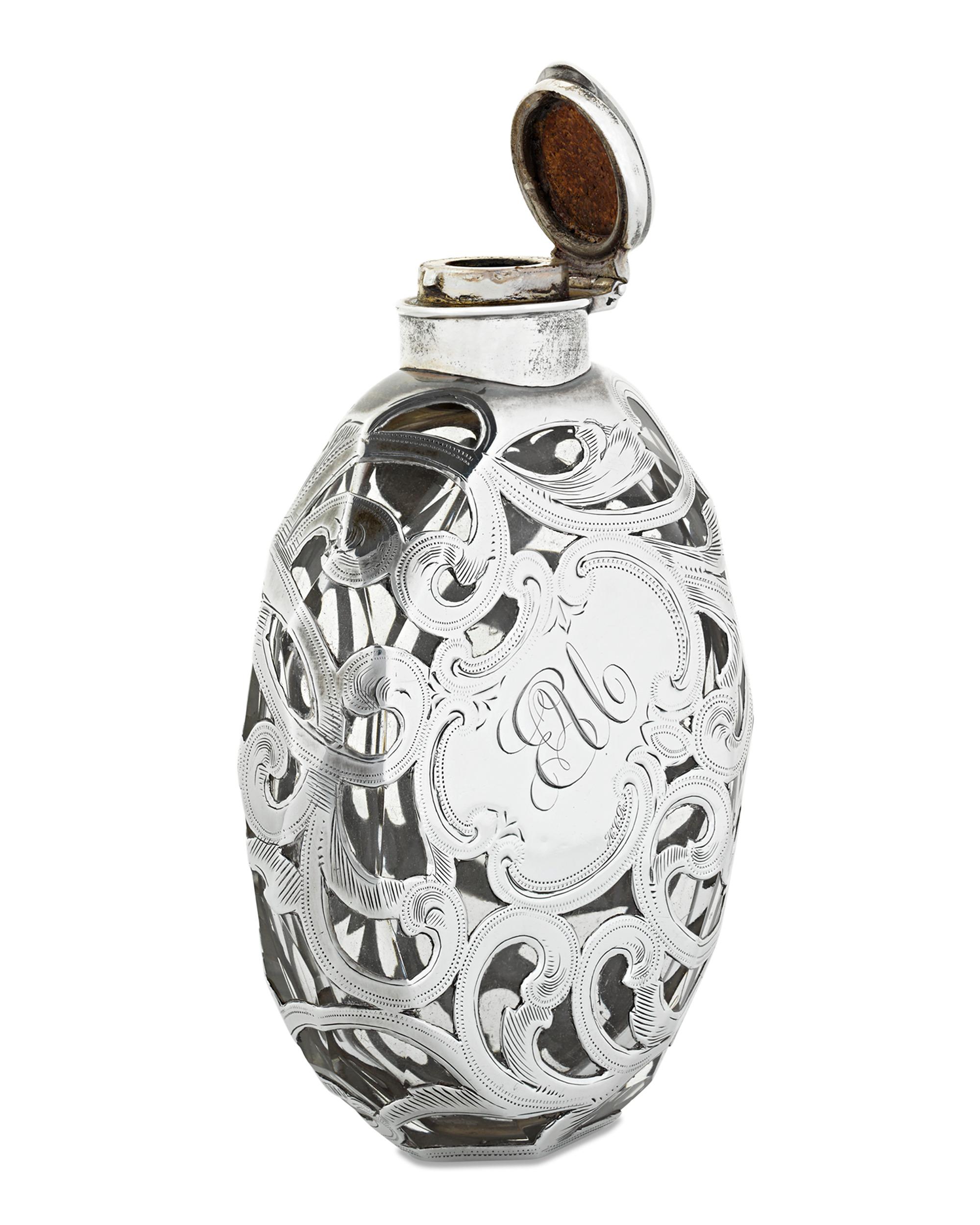 The elegance of rare silver overlay distinguishes this Art Nouveau glass flask. The flowing, graceful motif allows sections of the clear glass, and the contents within, to be visible. The sterling silver hinged lid secures the liquid