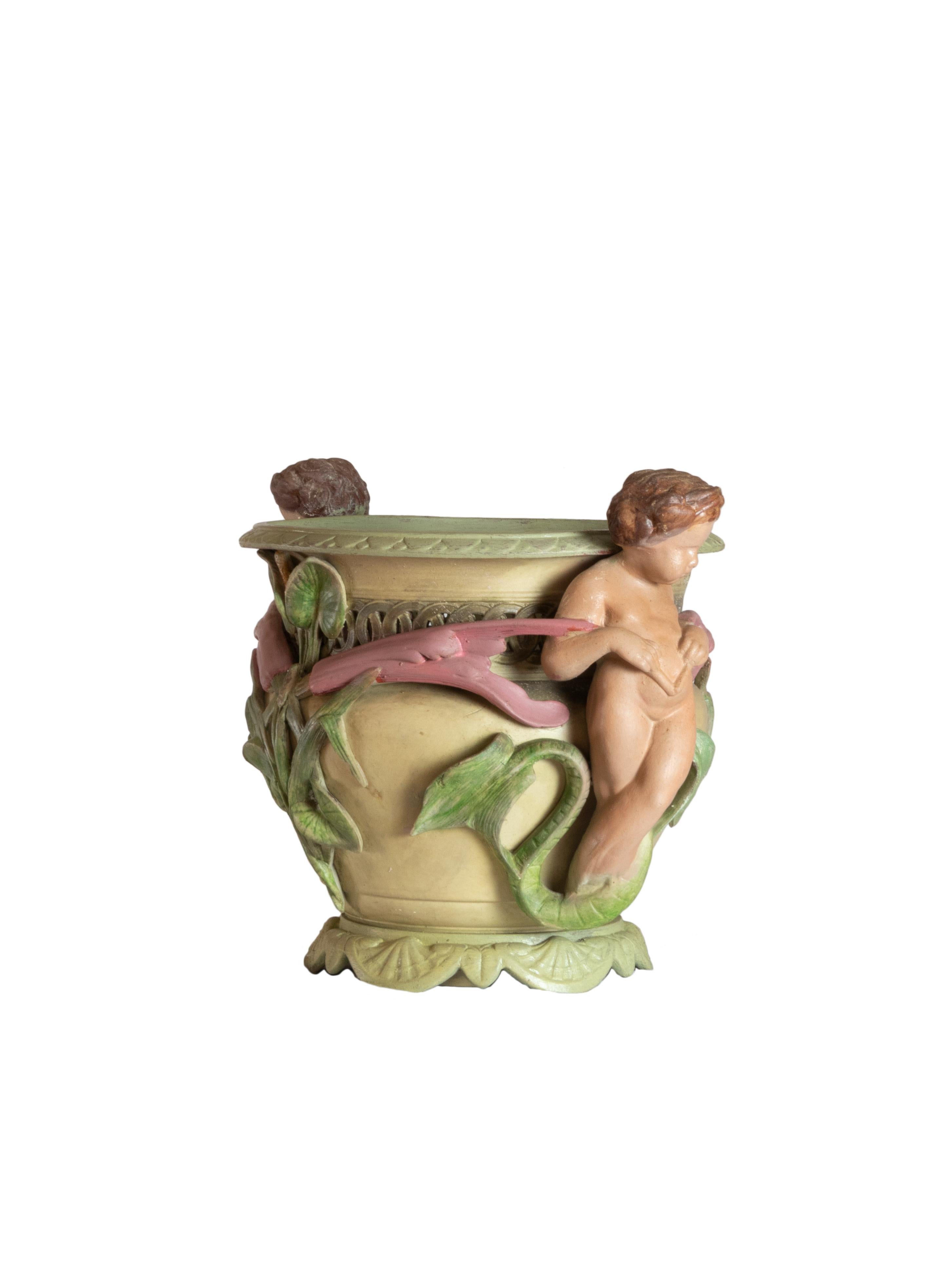 Una maceta de cerámica policromada presenta un diseño escultórico ornamentado con querubines en altorrelieve, entrelazados con motivos botánicos, como hojas y flores en flor. 

Los querubines flanquean el recipiente simétricamente, abrazando la