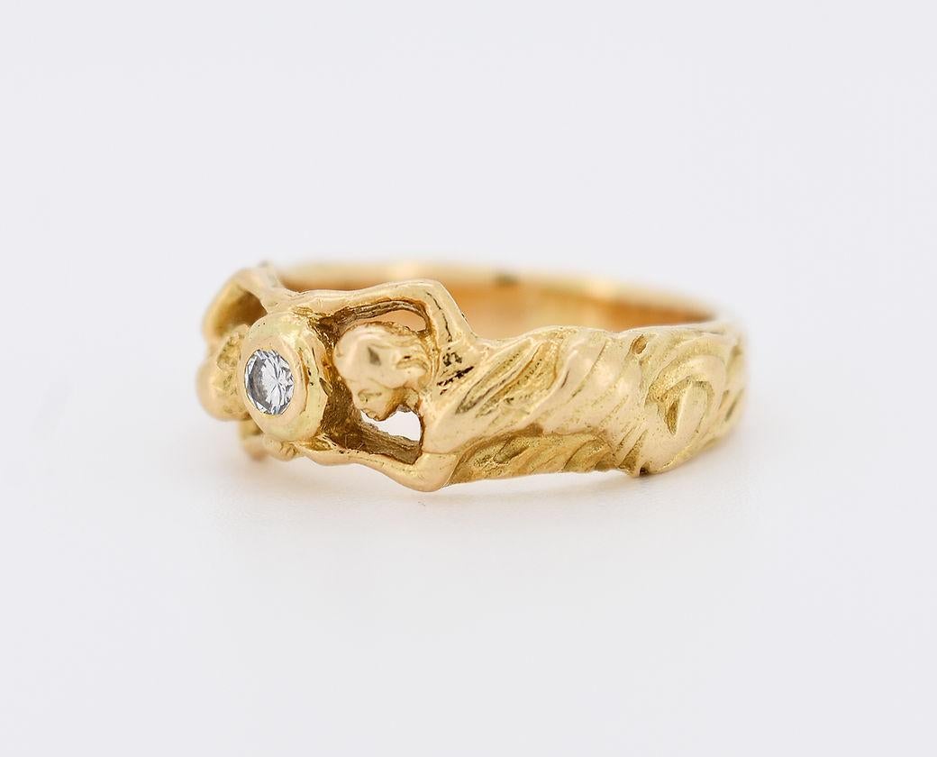 Art Nouveau ring in 18K gold (750/1000). It features a bezel-set, brilliant-cut central diamond of approximately 0.10 carats. The hand-crafted band showcases an exquisite design of two draped women intertwined around the diamond, exemplifying the