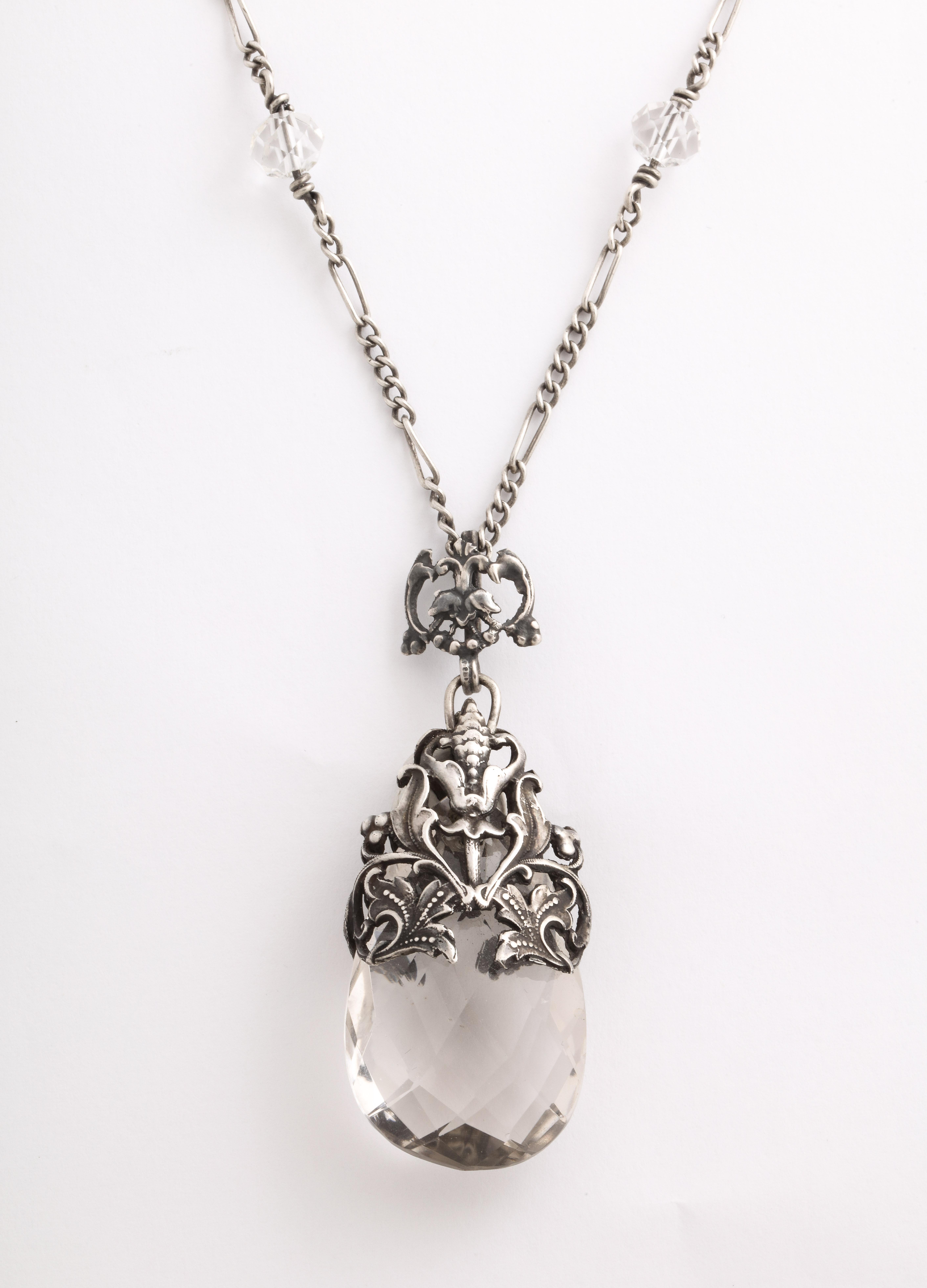 De style Art nouveau, ce collier en cristal de roche est magnifiquement conçu. Le cristal est gracieusement enveloppé dans des feuilles et des bourgeons gravés en acier inoxydable. Le cristal de roche est facetté en diagonale. La chaîne est
