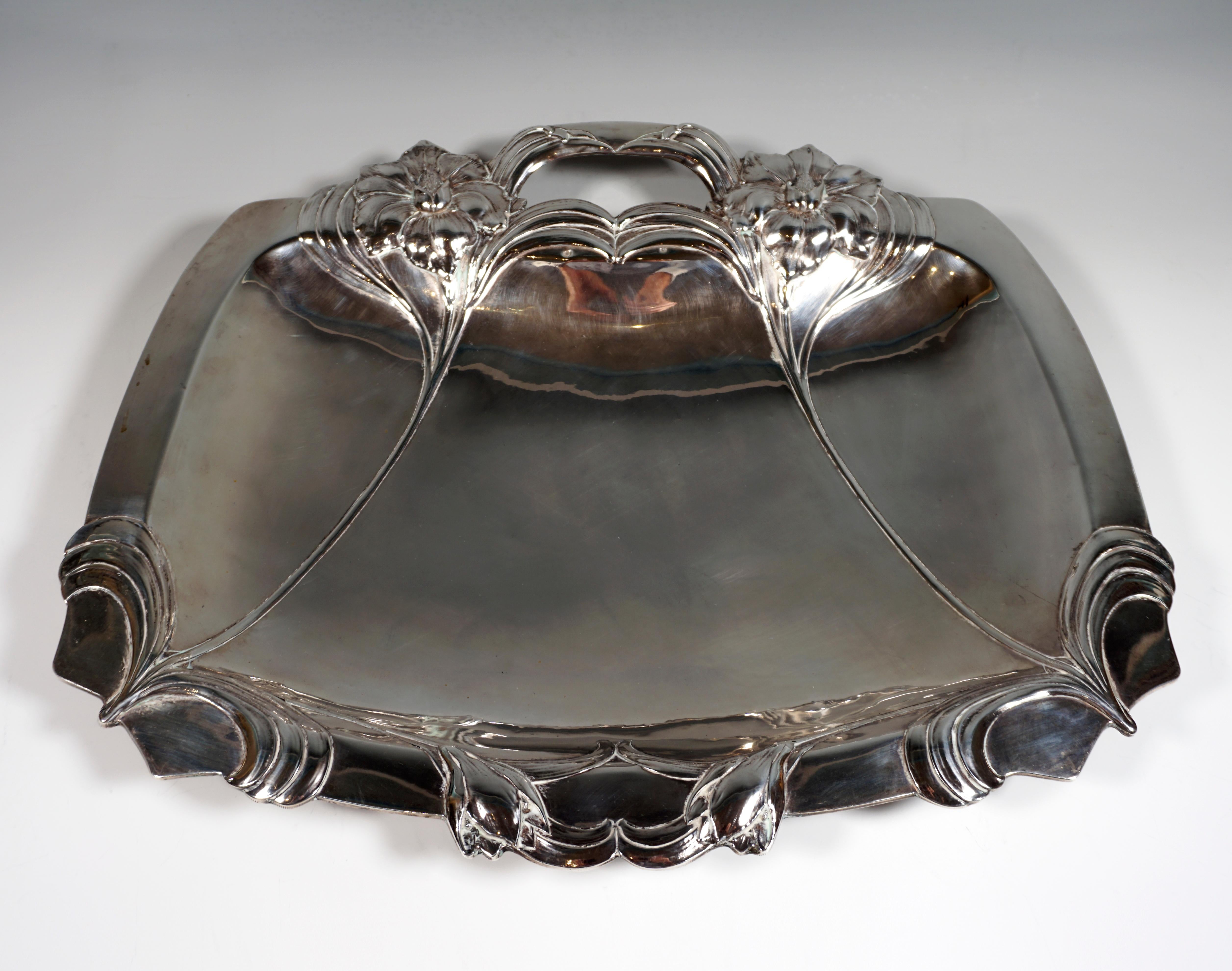 Wide silver bowl in a trapezoidal shape standing on four appliquéd feet, one-sided handle in the extension of two relief flower stalks with flowers and stylized leaves that arise in the opposite, rounded corners, irregular edge due to the flowers