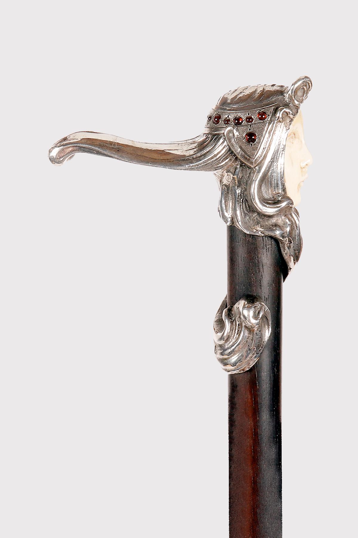 Bastón Art Nouveau. El pomo de plata representa el rostro de una niña, de marfil tallado, con largos cabellos enrollados a lo largo del cañón de madera de ébano. El peinado de la niña lleva piedras, granates cabujón, a lo largo de la banda del