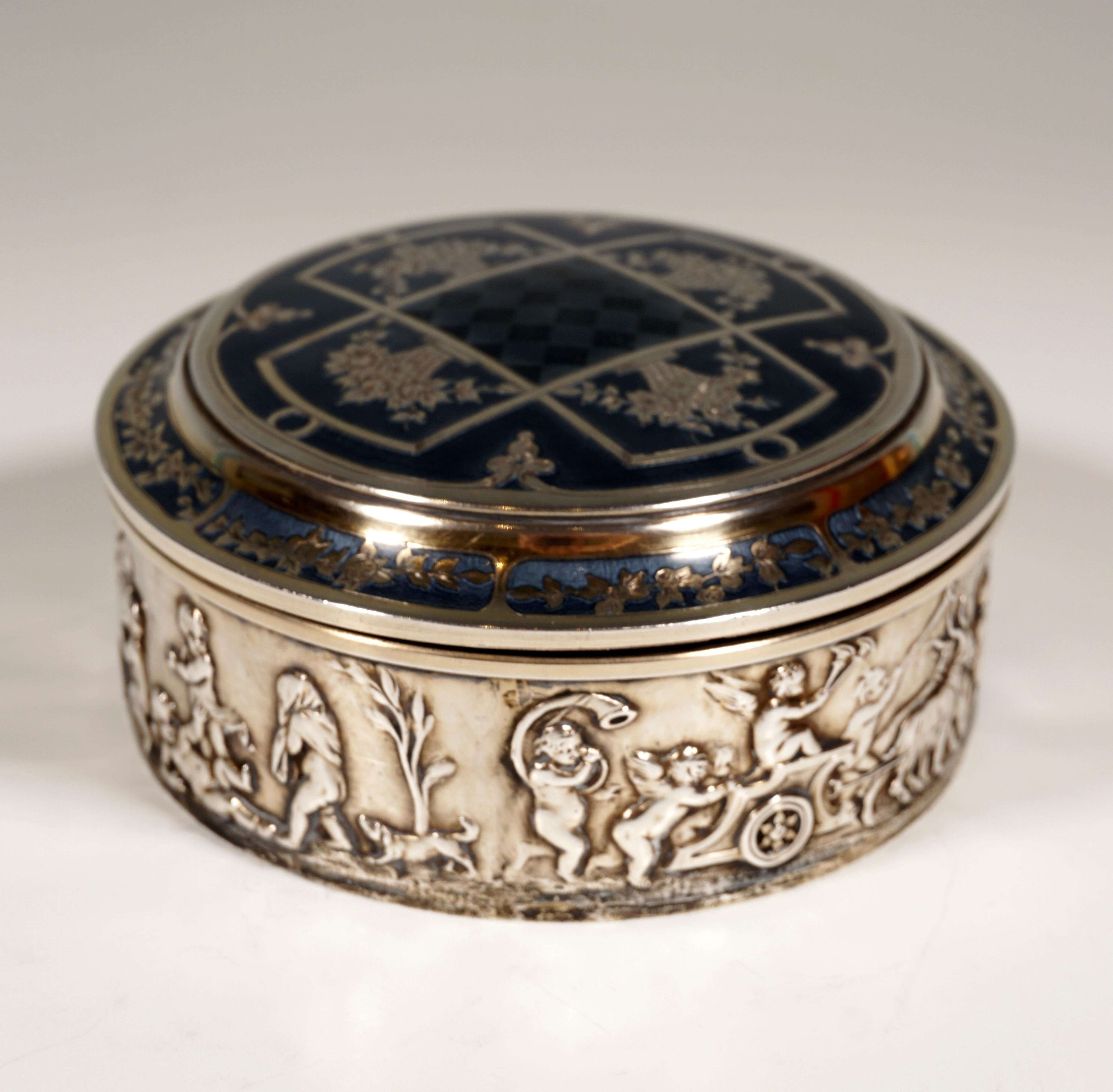 Cylindrical silver box with a circumferential relief band depicting a procession of playing and music-making putti and cupids, hinged lid with grooved edges and slightly arched in the middle with chased flower bouquet 
and tendril decoration,