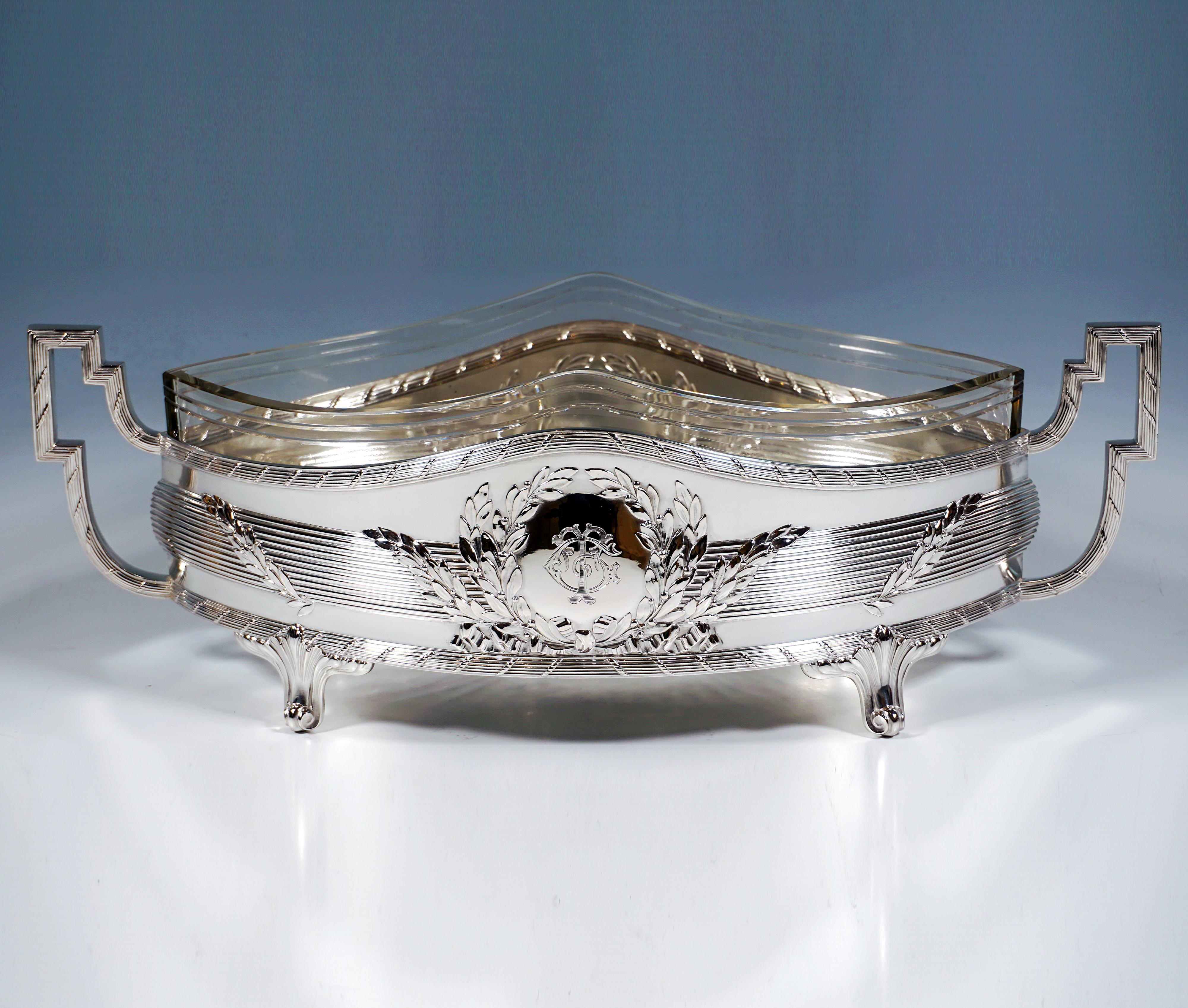 Elegante jardinera de plata en forma de barquito que se alza sobre cuatro pies de rocaille que sobresalen hacia abajo, el recipiente en la parte central con una banda horizontal de lamelas que lo rodea y se curva ligeramente hacia fuera, rodeada por