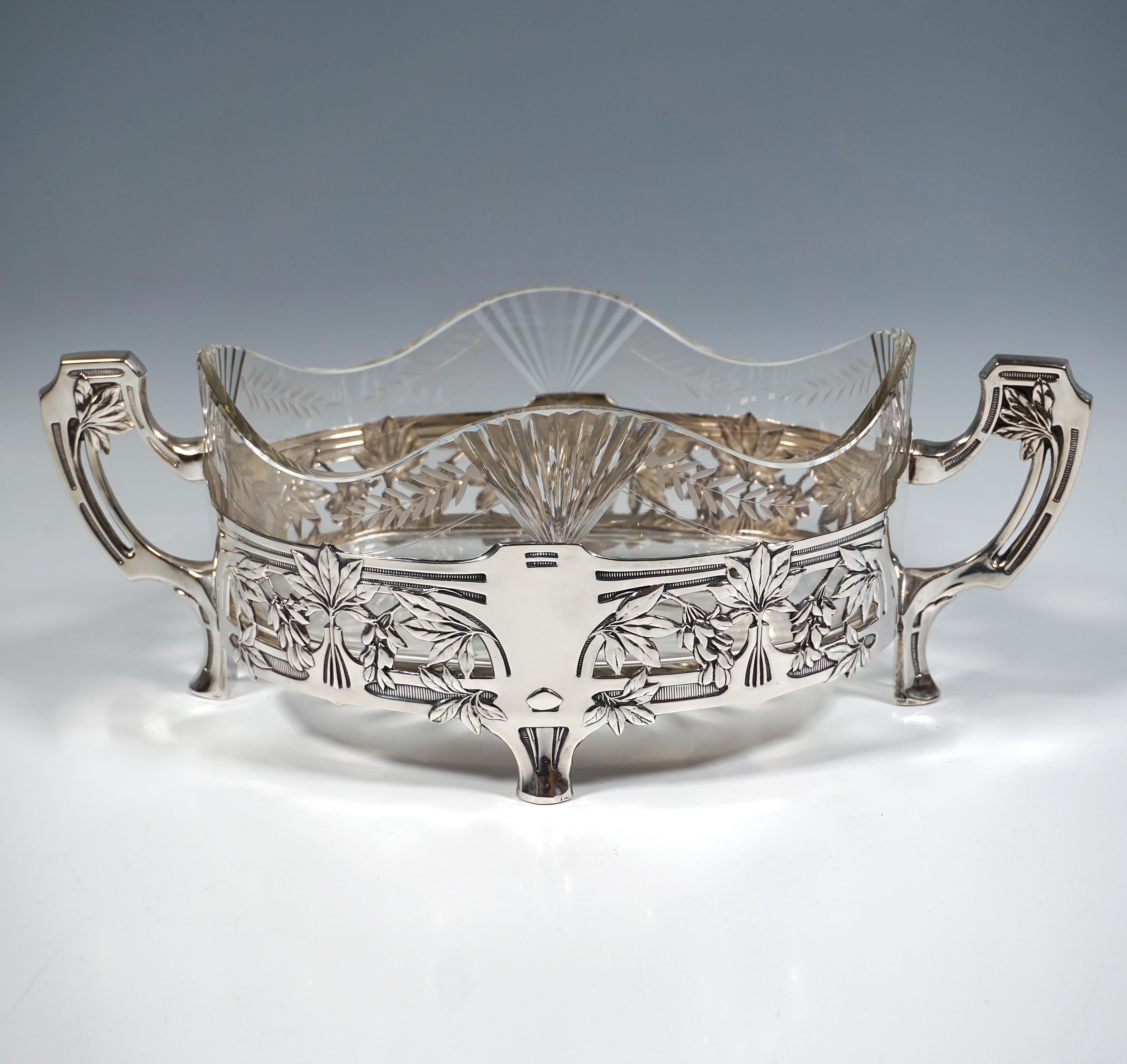 Elegante y festivo centro de mesa de plata con cuerpo ovalado sobre cuatro pies en voladizo con decoración en relieve, pared vertical con decoración floral calada en relieve en forma de cuatro ventanas de arco plano rellenas de follaje, borde