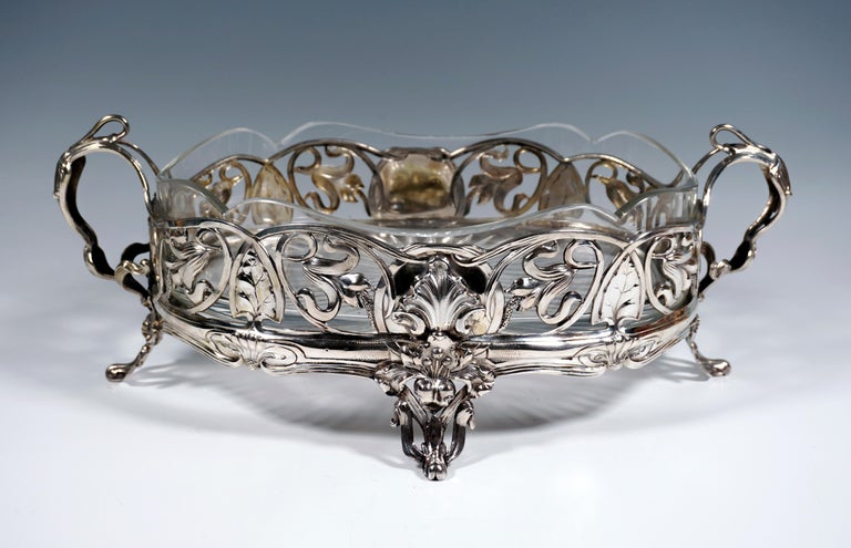 Silver vessel in an oval basic shape standing on four feet, completely openwork, with depictions of intertwined ribbons and tendrils, filled with flower and leaf motifs, two raised openwork handles with floral decoration.
Original glass insert with