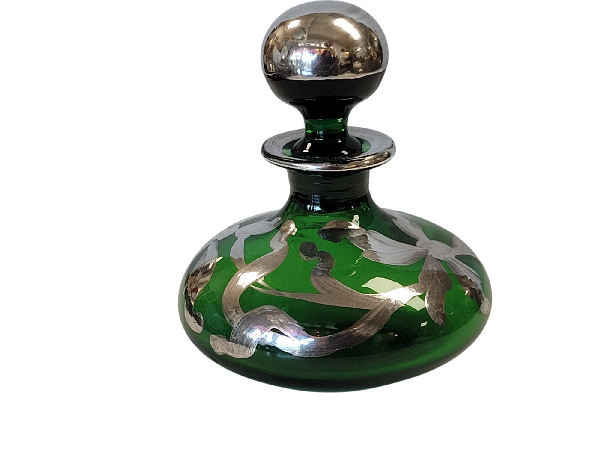 A refined example of late 19th to early 20th-century vanity art, this decorative perfume bottle features a deep emerald green glass body with a narrow neck and fitted glass stopper. The surface is overlaid with a metallic paint finish, often