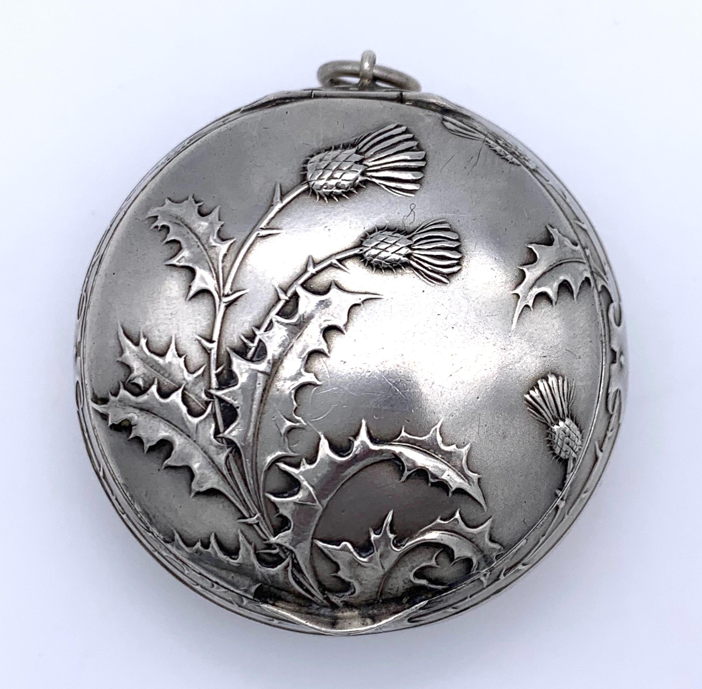 Anusual Art nouveau round silver box to be worn as a pendant. On all sides it is covered  with embossed thistle flowers and leaves. The inside of the box has been guilded.  