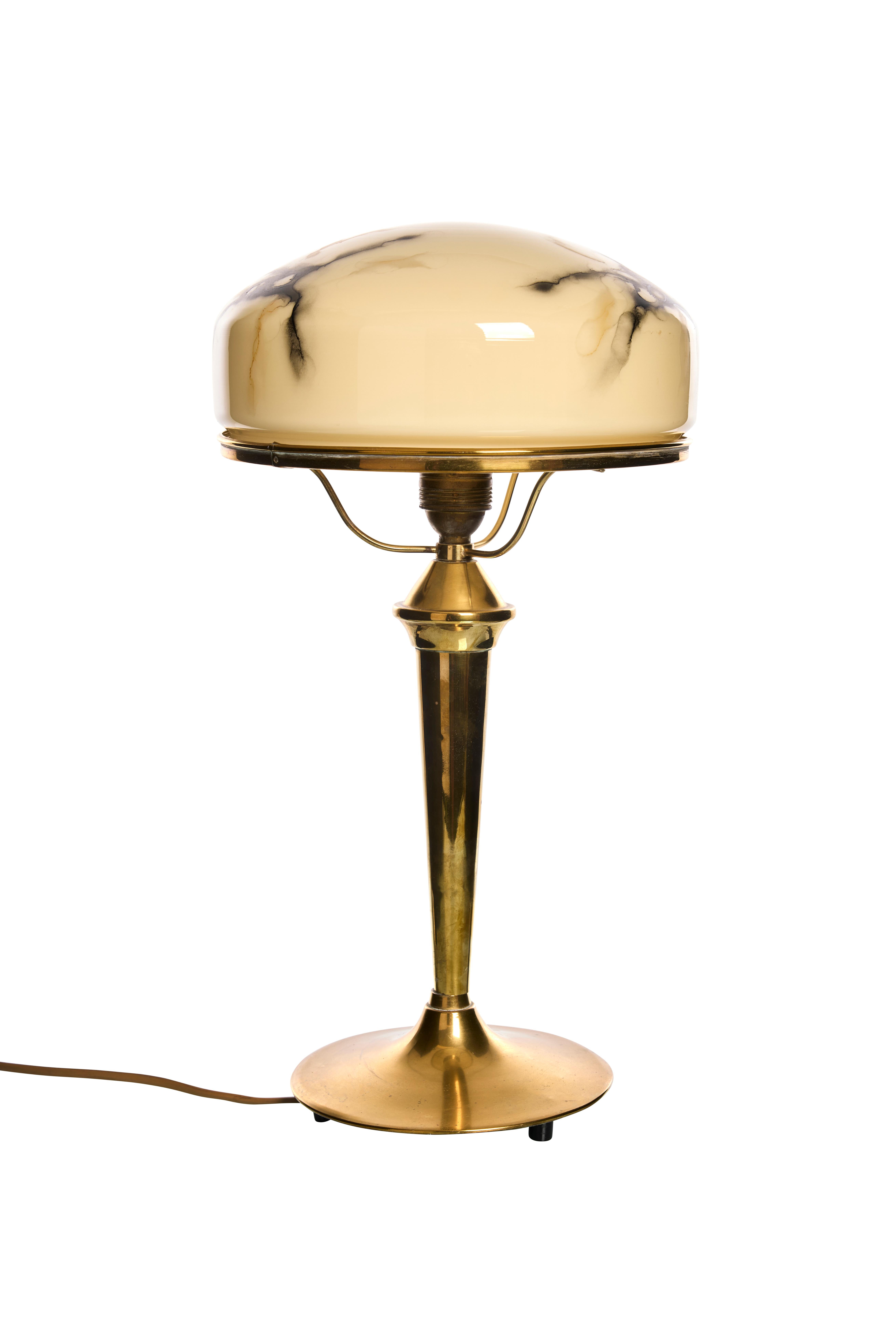 This table lamp features a brass base with a tapered, streamlined column that provides a sturdy foundation. Topping the lamp is a unique dome-shaped glass shade in a warm beige hue, adorned with organic, smoky black marbling, giving it a soft,