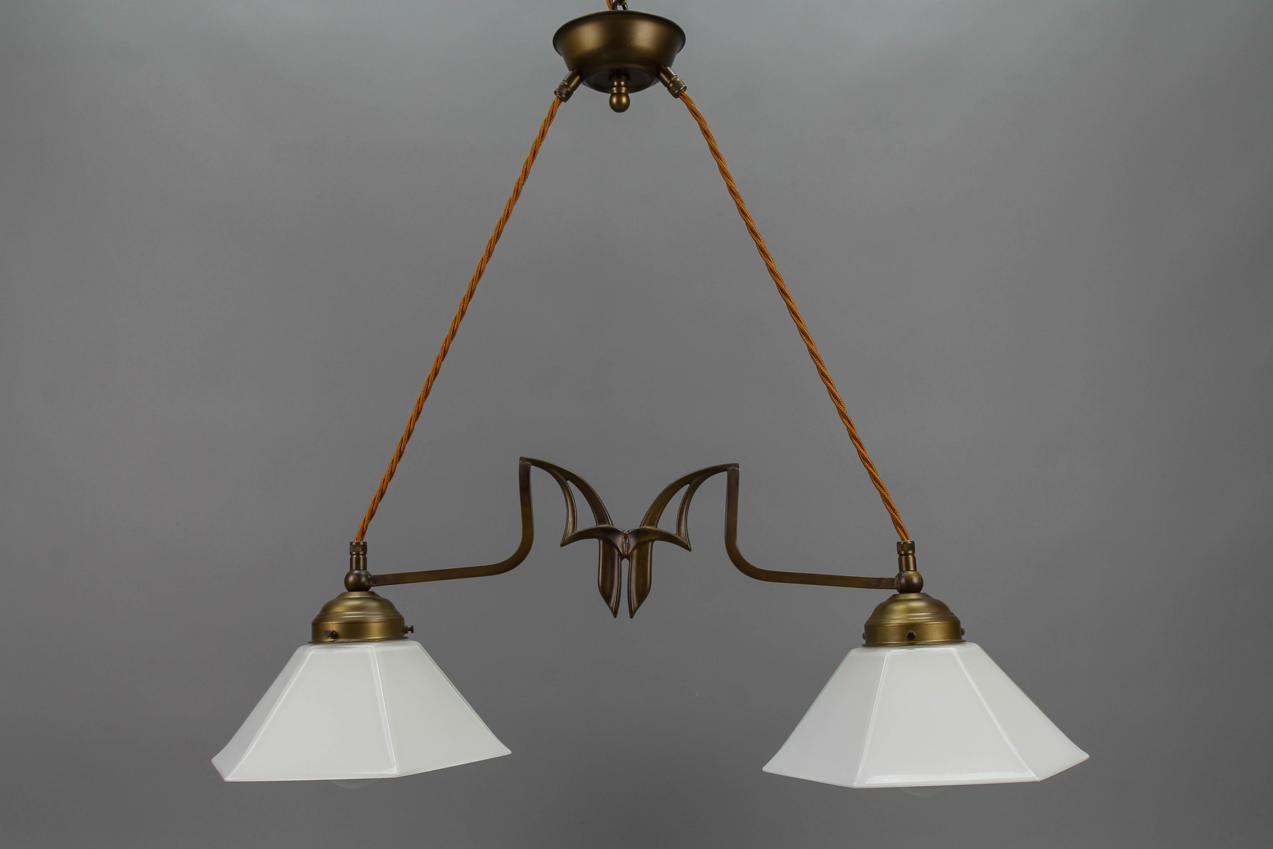 Art Nouveau style brass and white glass two-light pendant chandelier, Germany, circa the 1950s.
This elegant Art Nouveau-style pendant light fixture features two white glass hexagonal lampshades and a brass frame with a refined Art Nouveau-style