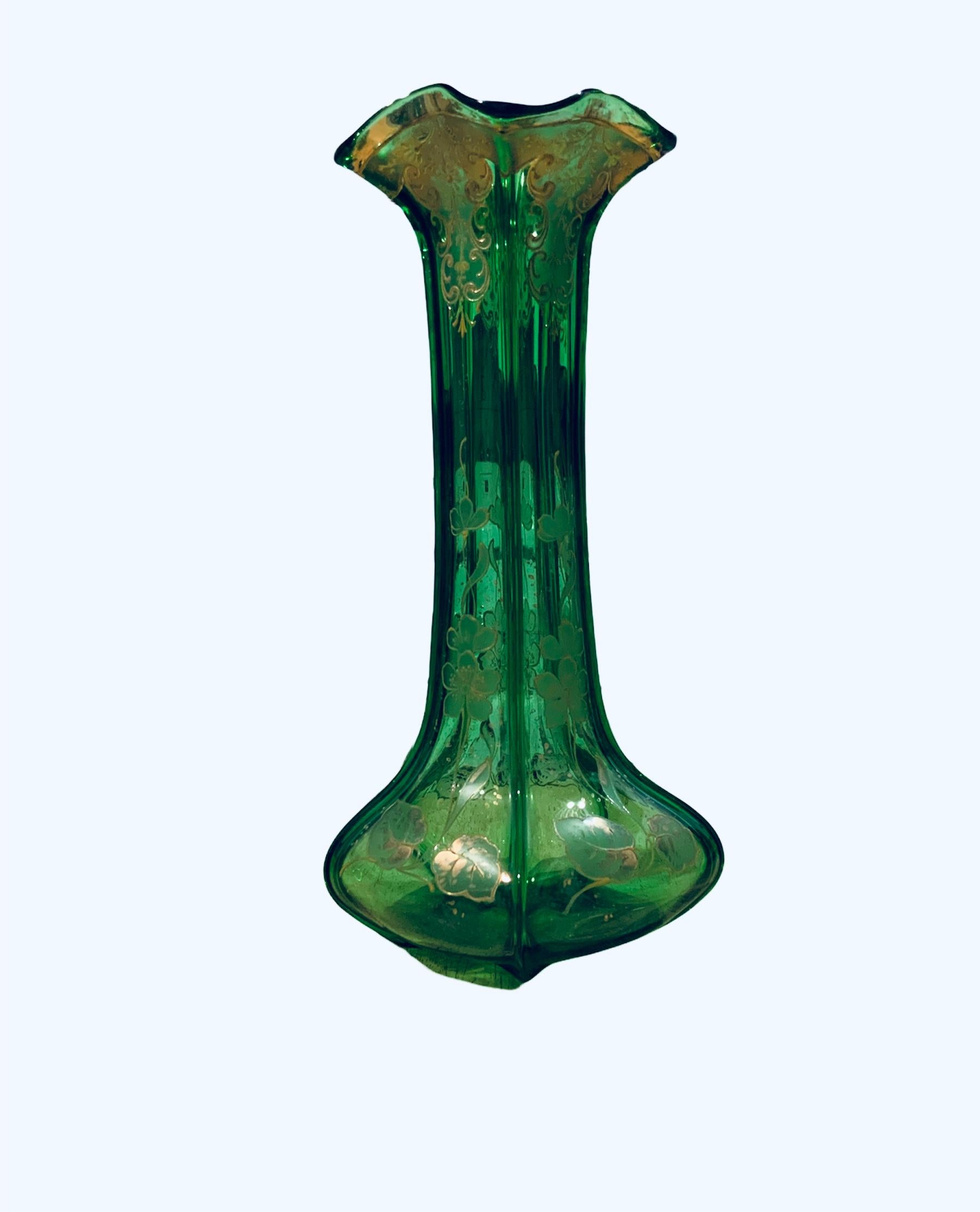 This is an Art Nouveau Style Gold enamel emerald green glass long vase. It depicts a vase with a star shaped bulbous base, long reeded neck and ruffled rim. It is adorned with gold painted branches of leaves and violets flowers. Also, gold C-scrolls