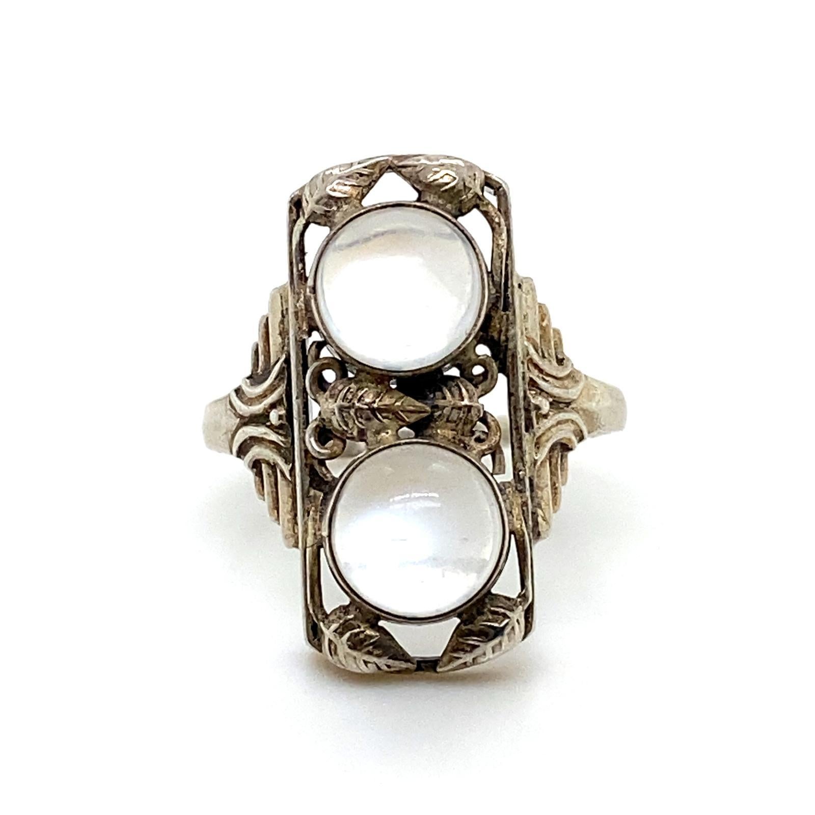 Ein silberner Mondstein-Plakettenring im Jugendstil, um 1920.

Dieser kühne Ring aus Silber hat eine längliche Form mit einem Laubsägearbeiten-Ausschnitt mit Blattmuster und ist mit zwei runden Cabochon-Mondsteinen besetzt.
Auf den Schultern