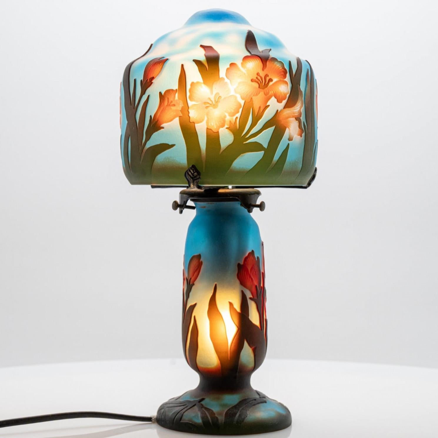 Esta hermosa lámpara es un ejemplo clásico del estilo Art Nouveau, conocido por sus formas elegantes y naturales. La lámpara está hecha de vidrio camafeo de varias capas, donde un detallado motivo floral en cálidos tonos naranja y rojo se revela