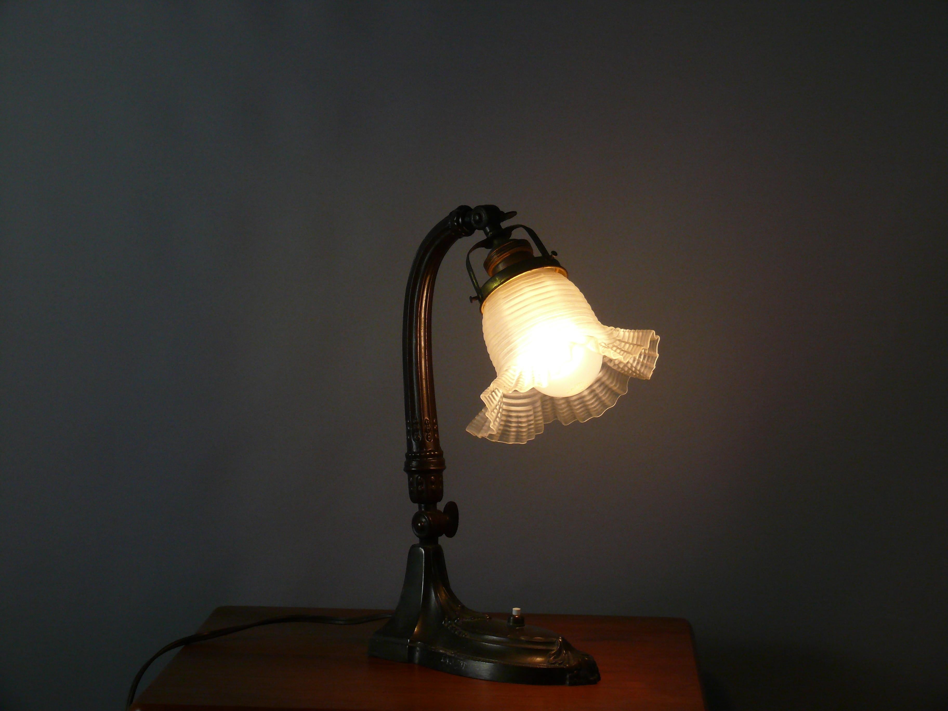Große originale Jugendstil-Tischlampe / Klavierlampe aus den 1910er Jahren in gutem Zustand. Die Leuchte ist aus Zinkdruckguss in Bronzeoptik gefertigt und hat einen blumenförmigen Glasschirm. Der Schirm ist in gutem Zustand. Sie kann über ein
