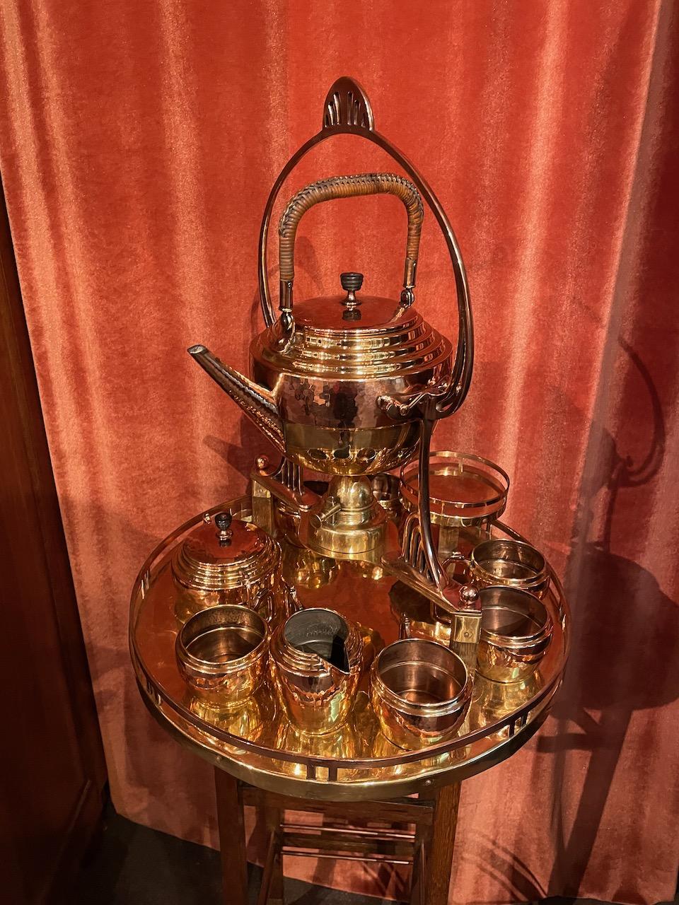 An Art Nouveau complete tea/coffee service in hand-hammered copper and brass on a graceful wooden stand. The set is comprised of a teapot with a woven wicker handle that hangs from a decorative metal arch over a brass chafing unit to keep the tea