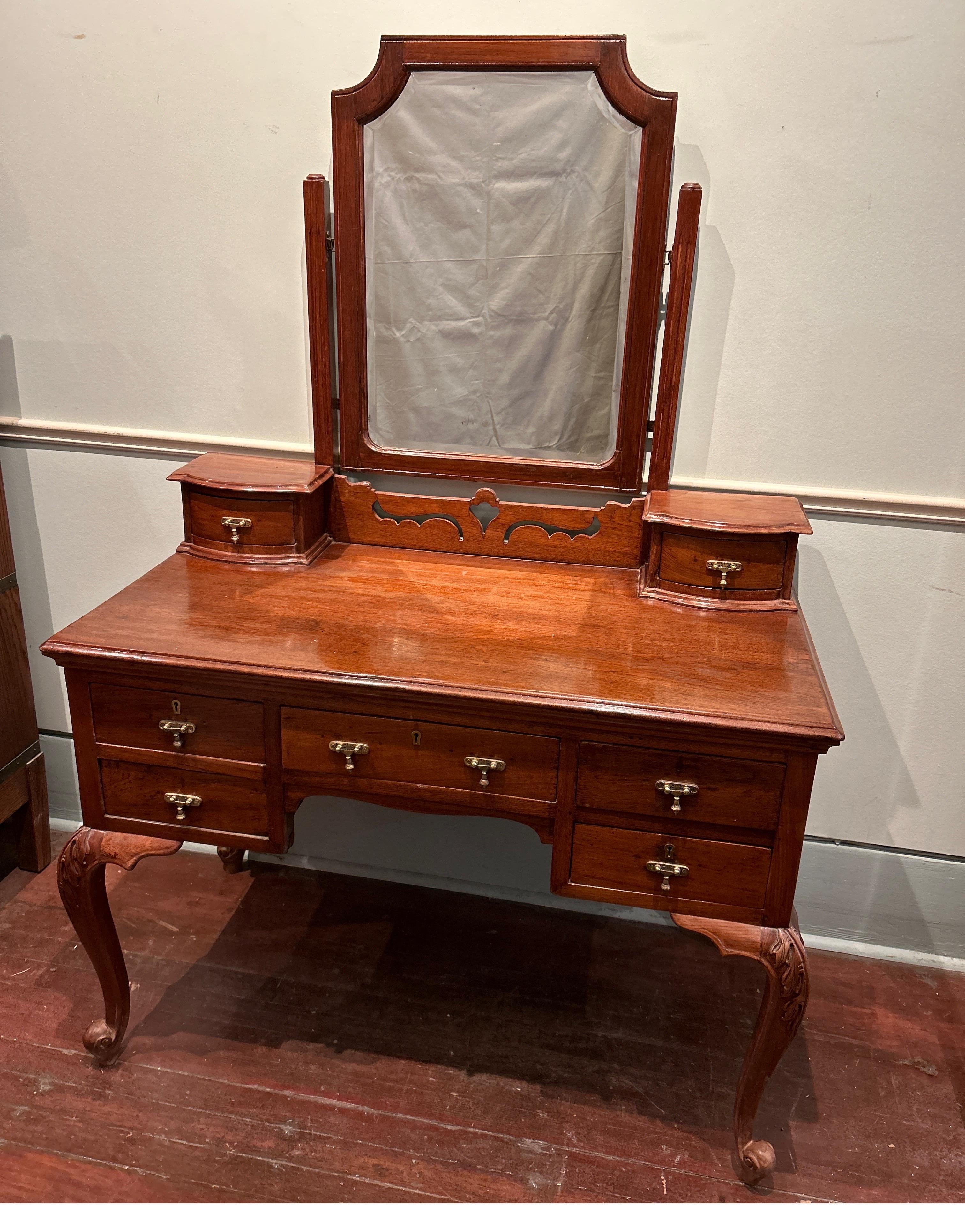 A vanity straight out the British Raj in Art Nouveau fashion. Solidly constructed from teak wood with hand carved scrolling legs and original bevel mirror and brass hardware. Nice panel work on the sides . Seven drawers make it very functional. A