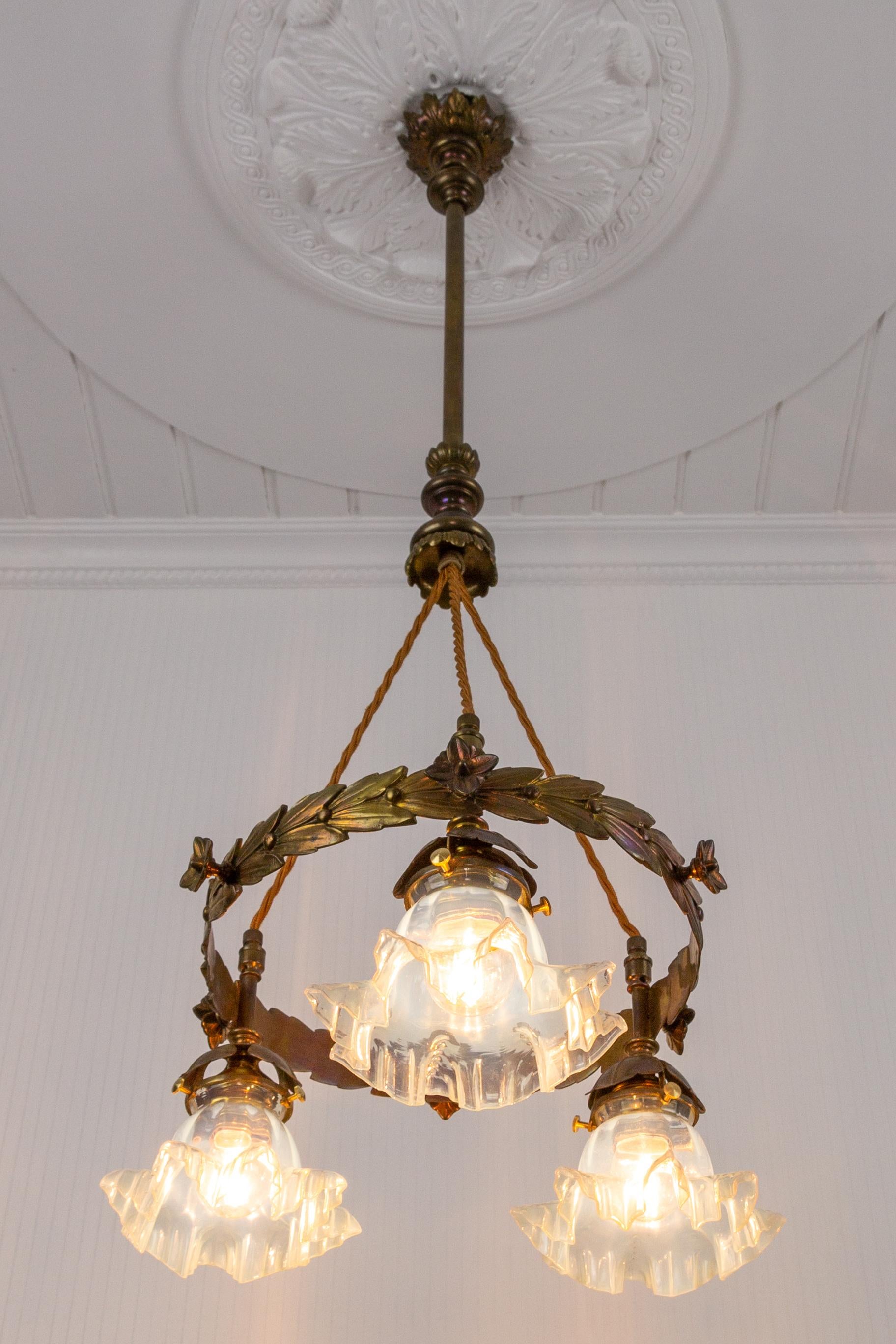 Beautiful Art Nouveau style brass chandelier from the 1920s. The pendant chandelier features brass parts with floral and acanthus leaves decorations and three floral-shaped lampshades made of opalescent translucent glass. 
Three sockets for E27