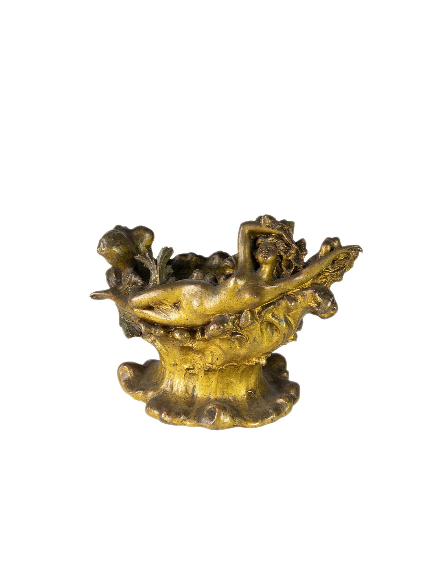 Un vaso molto particolare in metallo dorato con una statuetta di donna sdraiata riccamente decorata.
