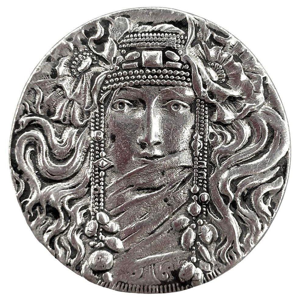 Broche/épingle en forme de portrait de femme voilée de Rine Pewter, datant du début du 20e siècle. Le portrait rappelle les portraits féminins peints par Alphonse Mucha.

La femme élégante est parée de tresses décoratives complexes et entourée de