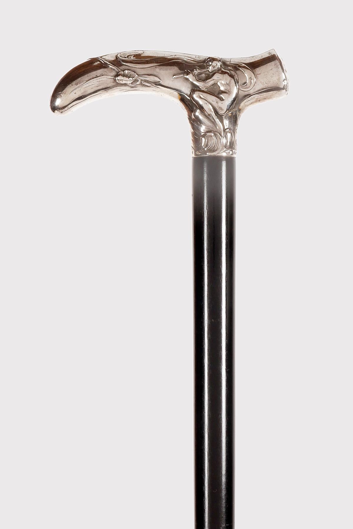 Canne Art Nouveau. Le bouton en argent 838/1000 en forme de T est estampé et ciselé et représente, d'un côté, le Dieu des bois, Pan, assis sur un rocher et jouant de la flûte, et de l'autre, la nymphe Echo jouant de la lyre. Manche en bois d'ébène.