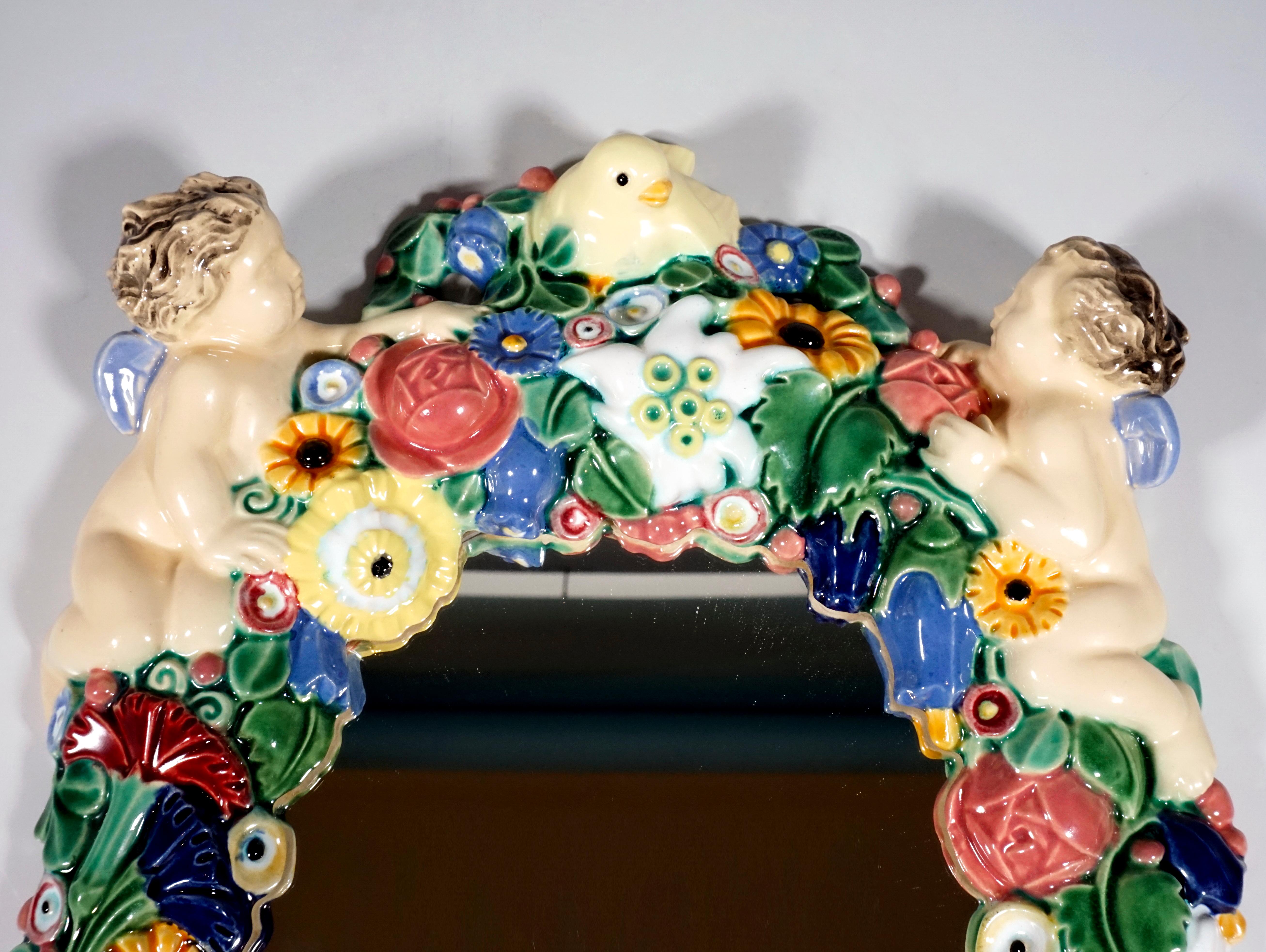 Oval mirror, framed by a dense, colored flower wreath made of ceramic, on the upper edge two winged putti, between them a bird in the middle.

By Michael Powolny (1871-1954)
Nowadays, art experts attest Michael Powolny’s work as the work of one