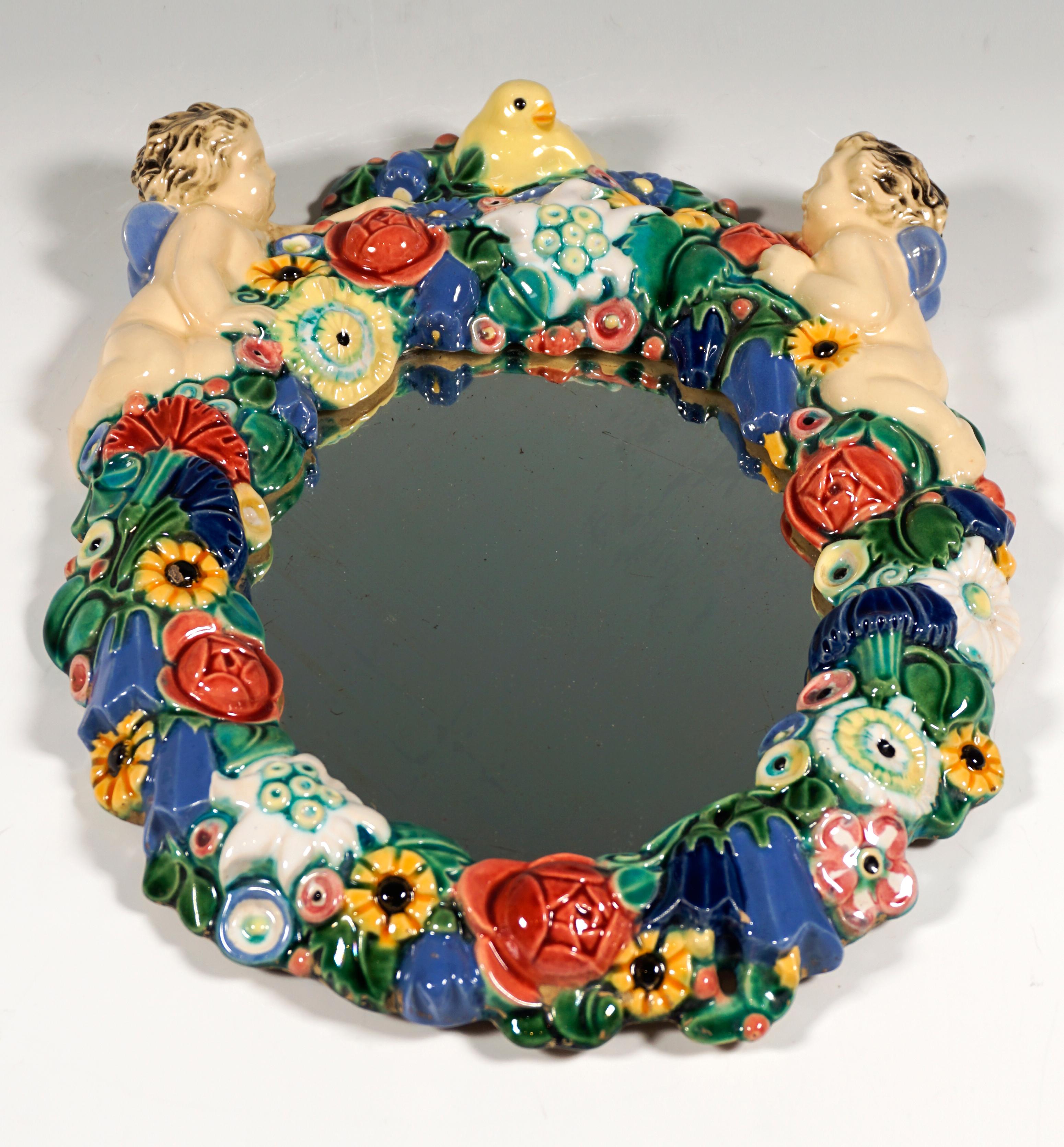 Oval mirror framed by a dense coloured floral wreath formed of ceramic, on the upper rim two winged putti, between them a bird in the middle.

By Michael Powolny (1871 - 1954)
Nowadays, art experts attest Michael Powolny’s work as the work of one of