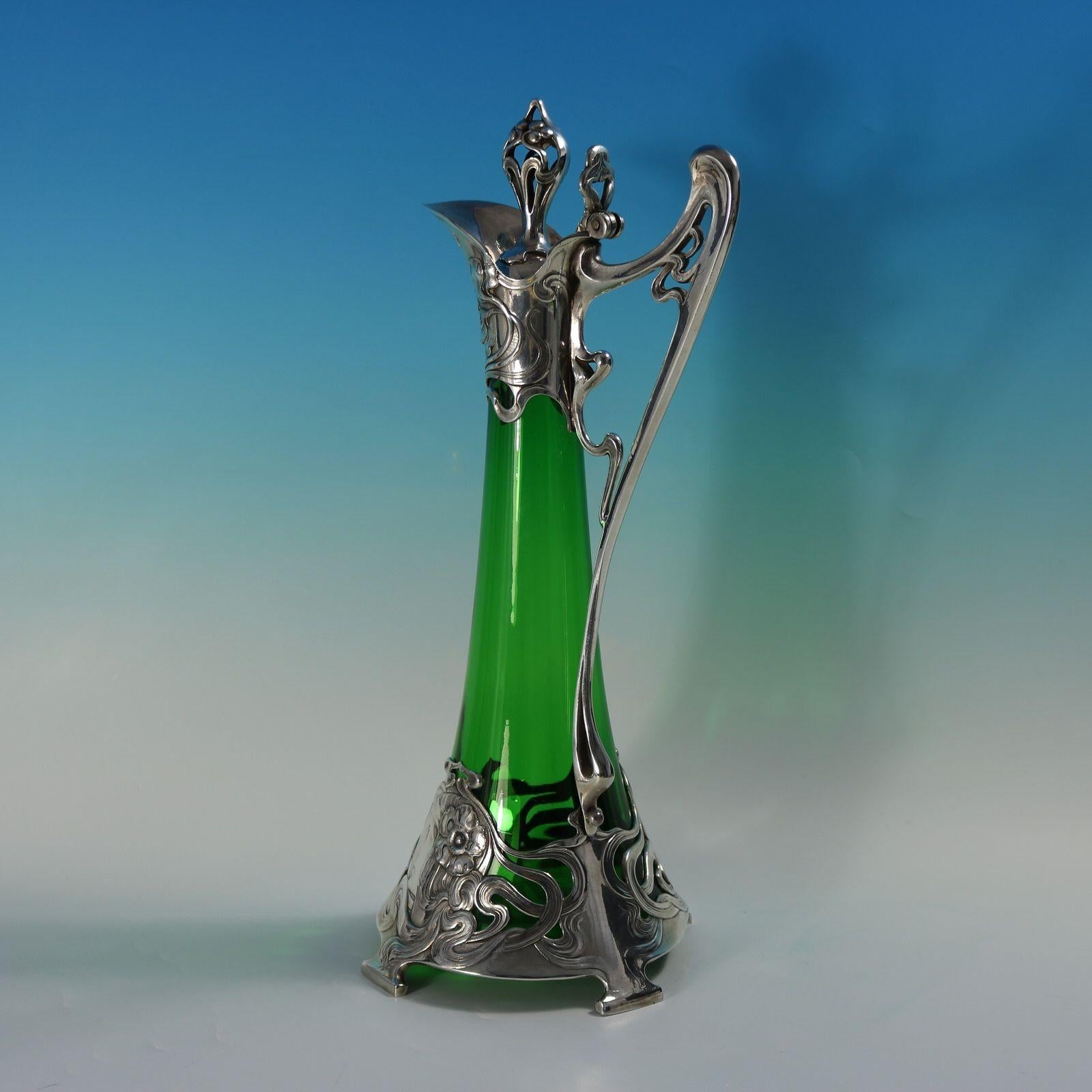 WMF green glass claret jug of typical art nouveau stylised form. The bottom mount depictis a maiden's head with a flower in her hair. The hair flows around the piece creating a winding pattern. The upper mount has floral motifs. The finial modelled
