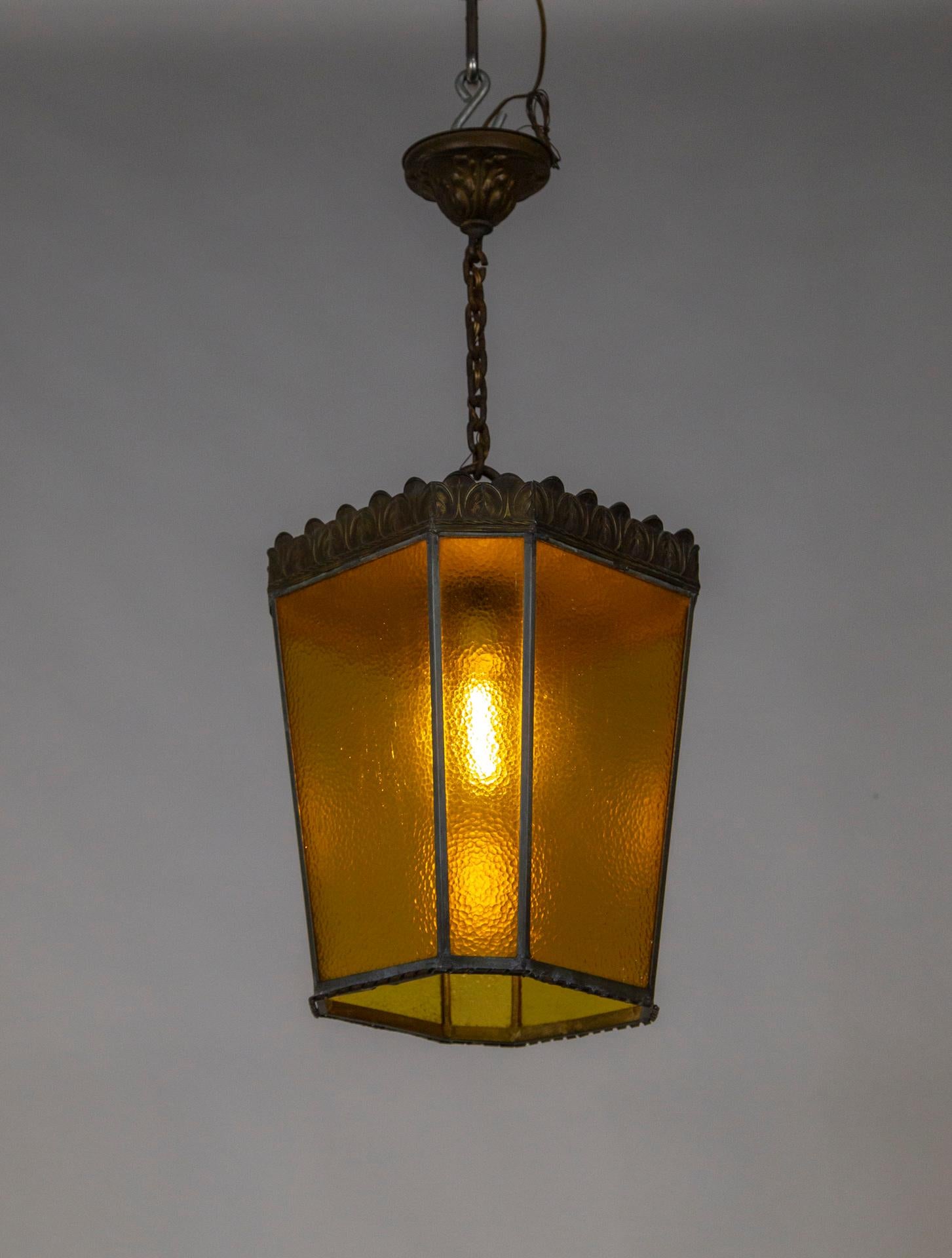 A large, early 20th-century lantern with 8 yellow, rippled glass panels (4 are wide and four narrow). Its brass armature has a dark, aged patina and is trimmed with imprinted, stylized leaves around the top, and a thin coil around the bottom. Newly