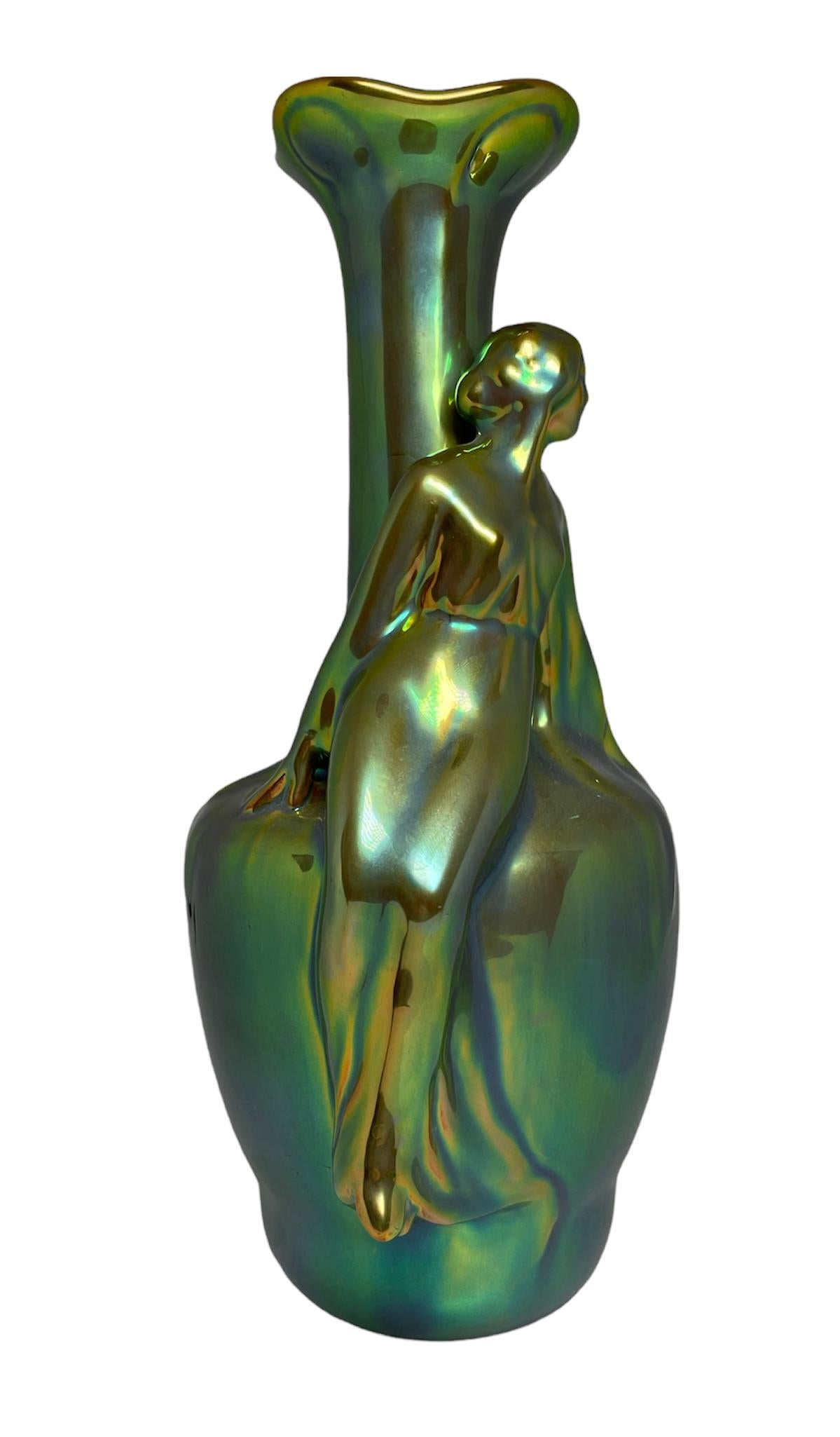 This is an Art Nouveau Zsolnay eosin green glazed ceramic vase. It depicts an urn shaped vase with long neck and a maid with a long dress looking to the side sat in the body of the urn with her back in from of its neck. The vase shows different