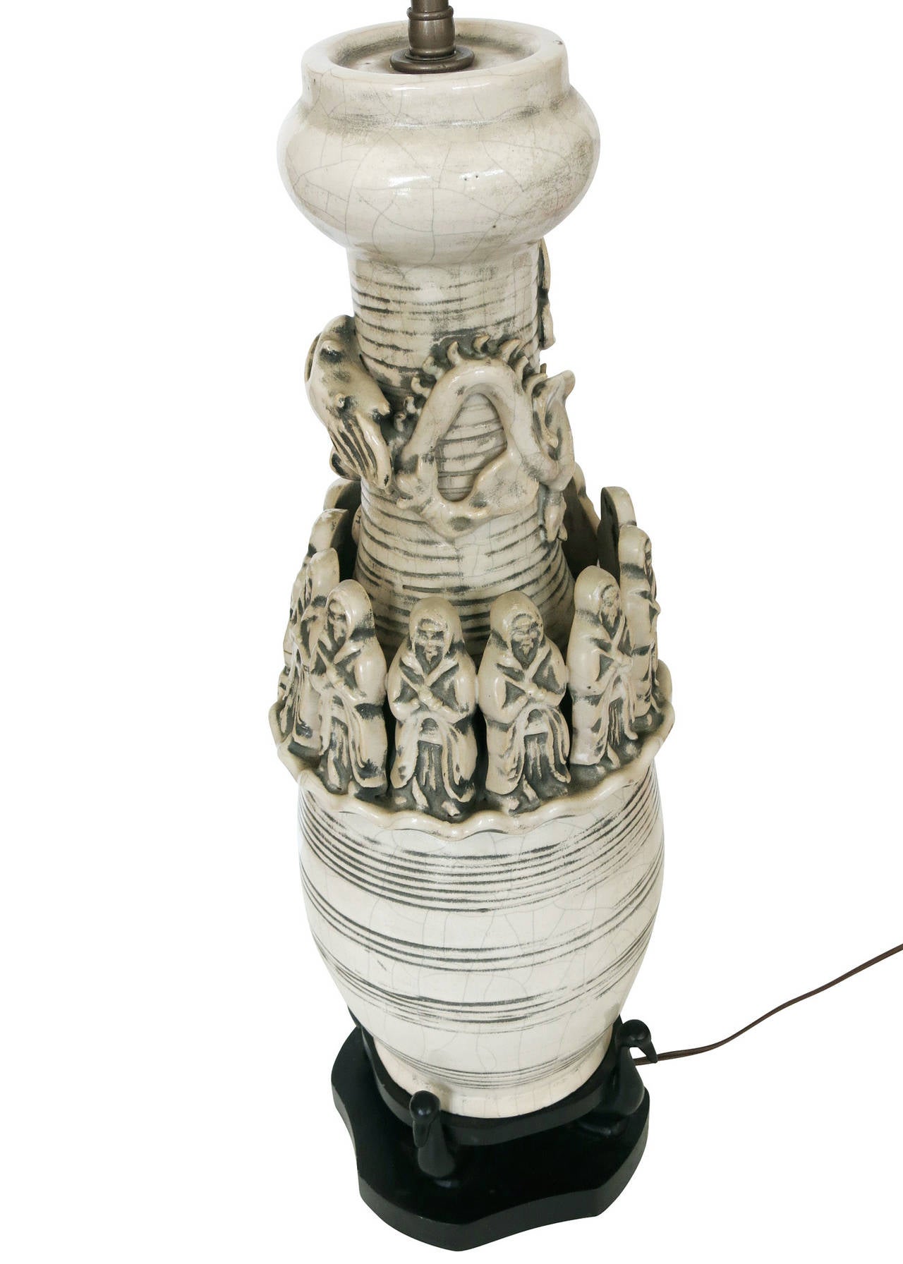 The work is a white art pottery lamp of a cylindrical shape with wood base. The lamp is decorated with 12 monks that surround a dragon. The lamp also has decorative, horizontal patterns and crackle finish. Carved out of the wood base that holds the