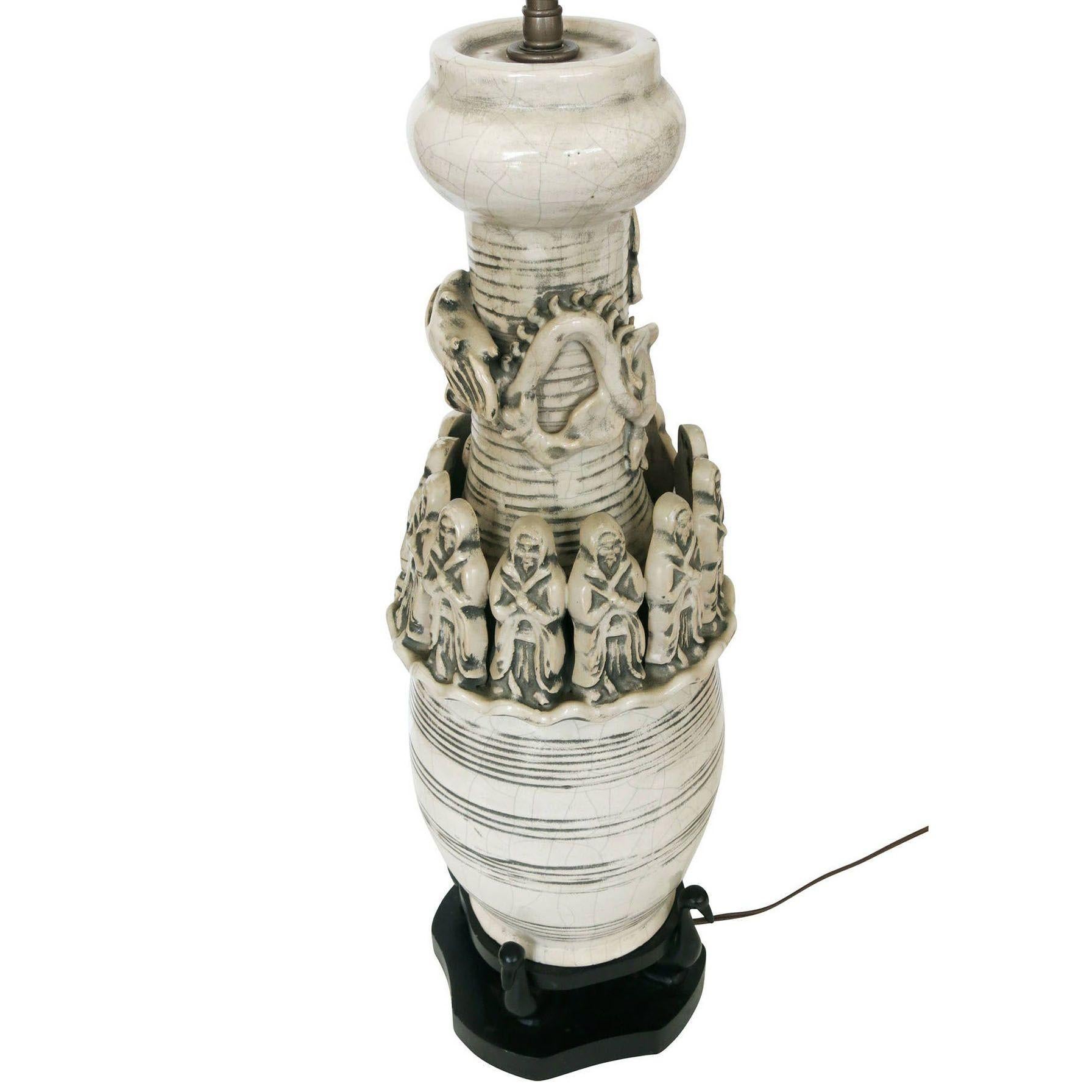 The work is a white art pottery lamp of a cylindrical shape with wood base. The lamp is decorated with 12 monks that surround a dragon. The lamp also has decorative, horizontal patterns and crackle finish. Carved out of the wood base that holds the