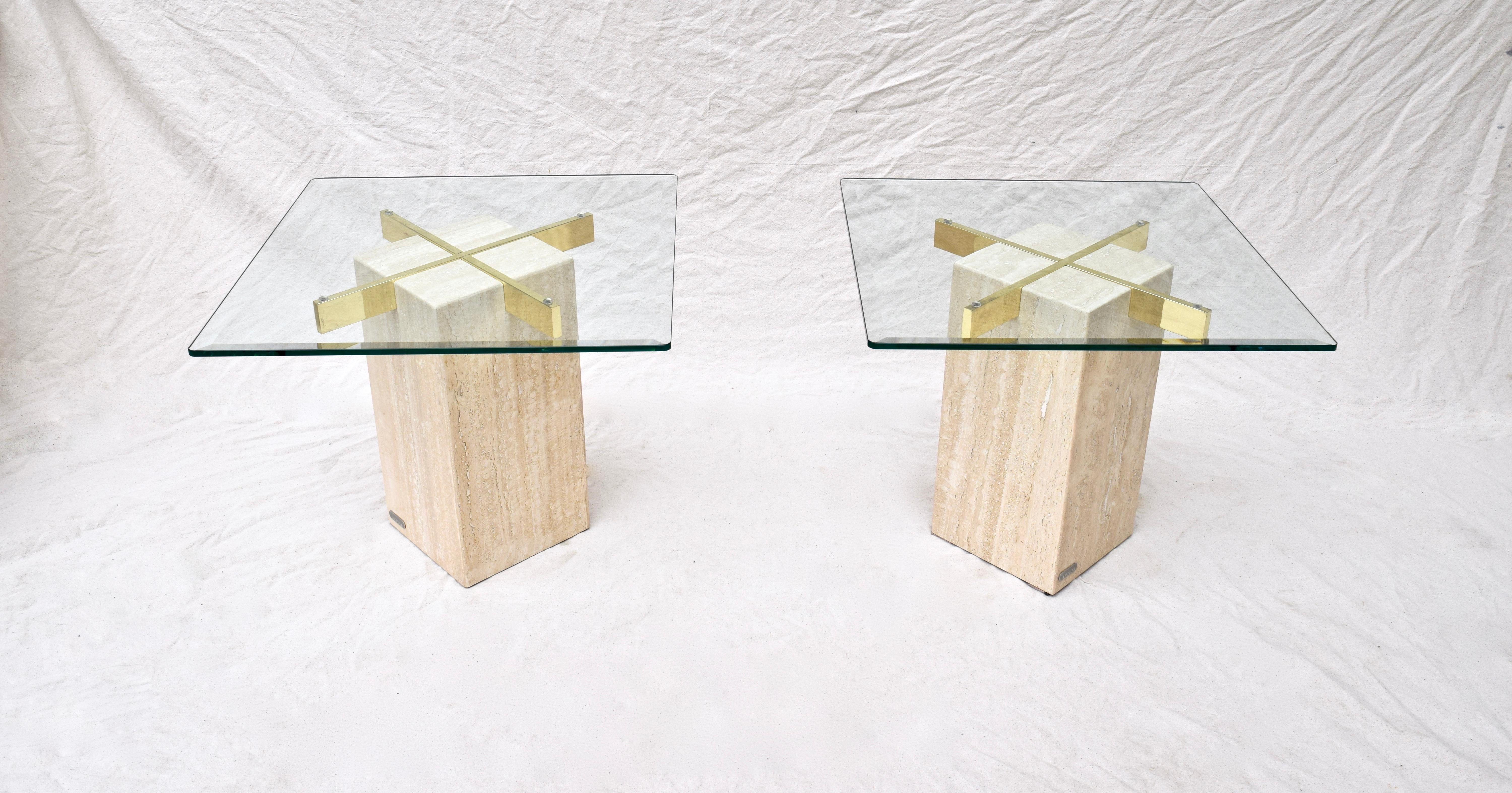 Paire de tables sculpturales fabriquées par Artedi au Royaume-Uni. Chaque table est dotée d'une base en marbre travertin, d'un plateau en verre subtilement biseauté et d'un support en X inversé en laiton poli. Les médaillons métalliques originaux