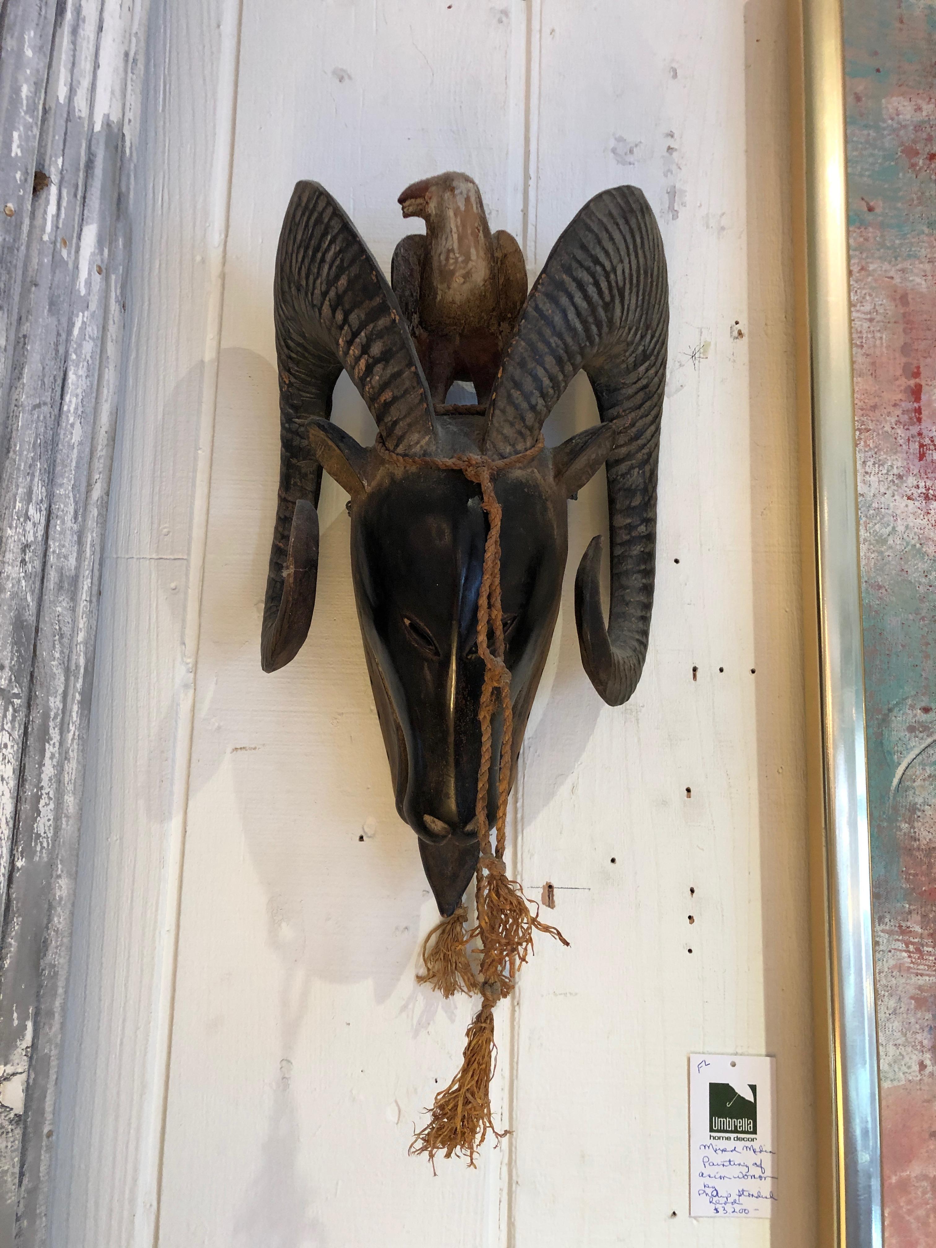 A striking wall sculpture from Africa that's a tribal mask of a stylized ram having smooth face with slanted eye holes and curly horns. A carved wood bird rests atop the head looking out between the horns.