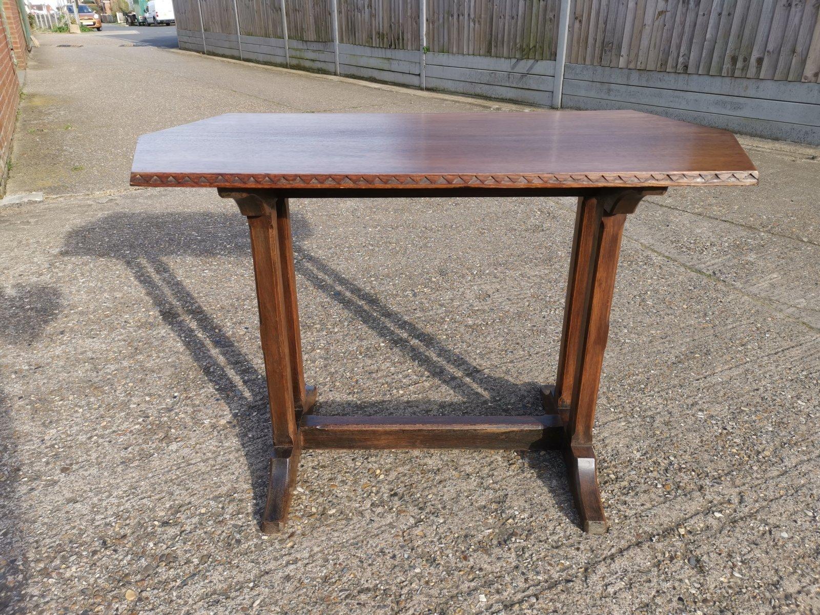 A Cotswold-style oak refectory dining or library table with carved chevron detailing to the edges of the double-angled ended top. On twin upright legs with 45 degree chamfered edges that unite to the shaped feet, the lower stretcher full length with