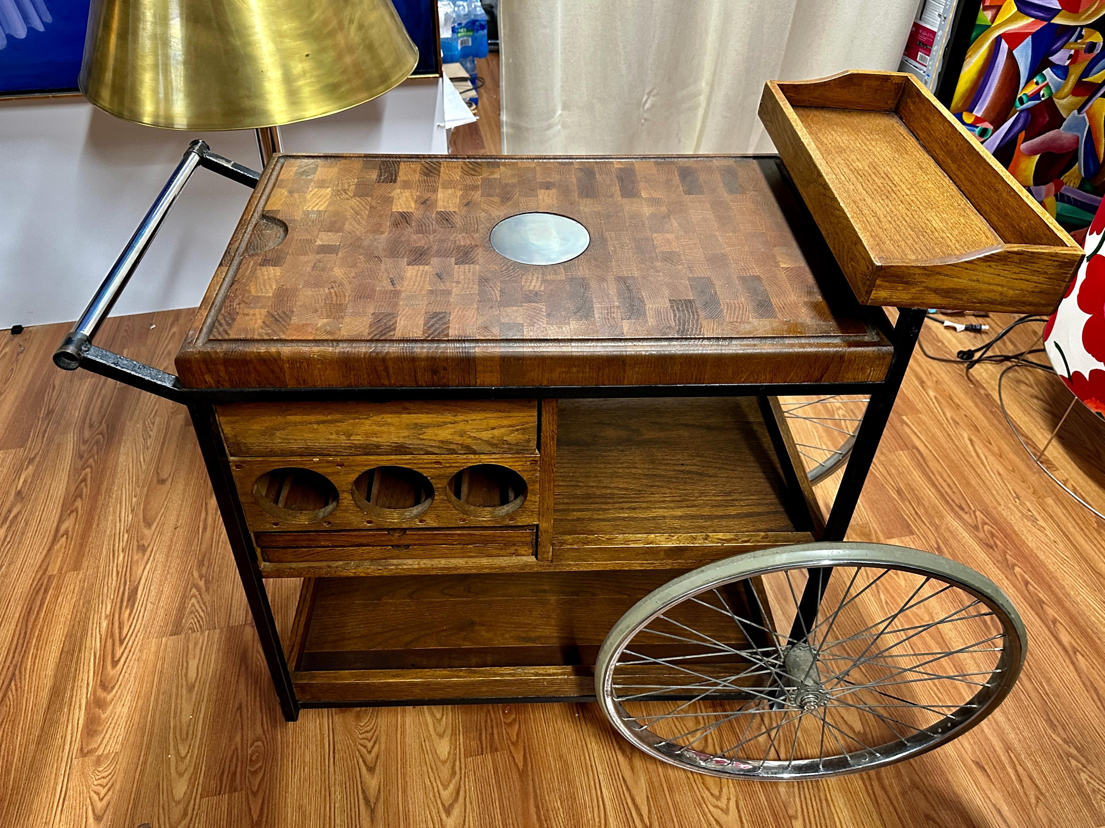 Great looking mid-century bar cart designed by Arthur Umanoff. Distinctive large pair of Rear wheels and a small front wheel for “steering.” Thick Butcher block top with oak trays and sides. The frame is painted black iron. An unusual design by