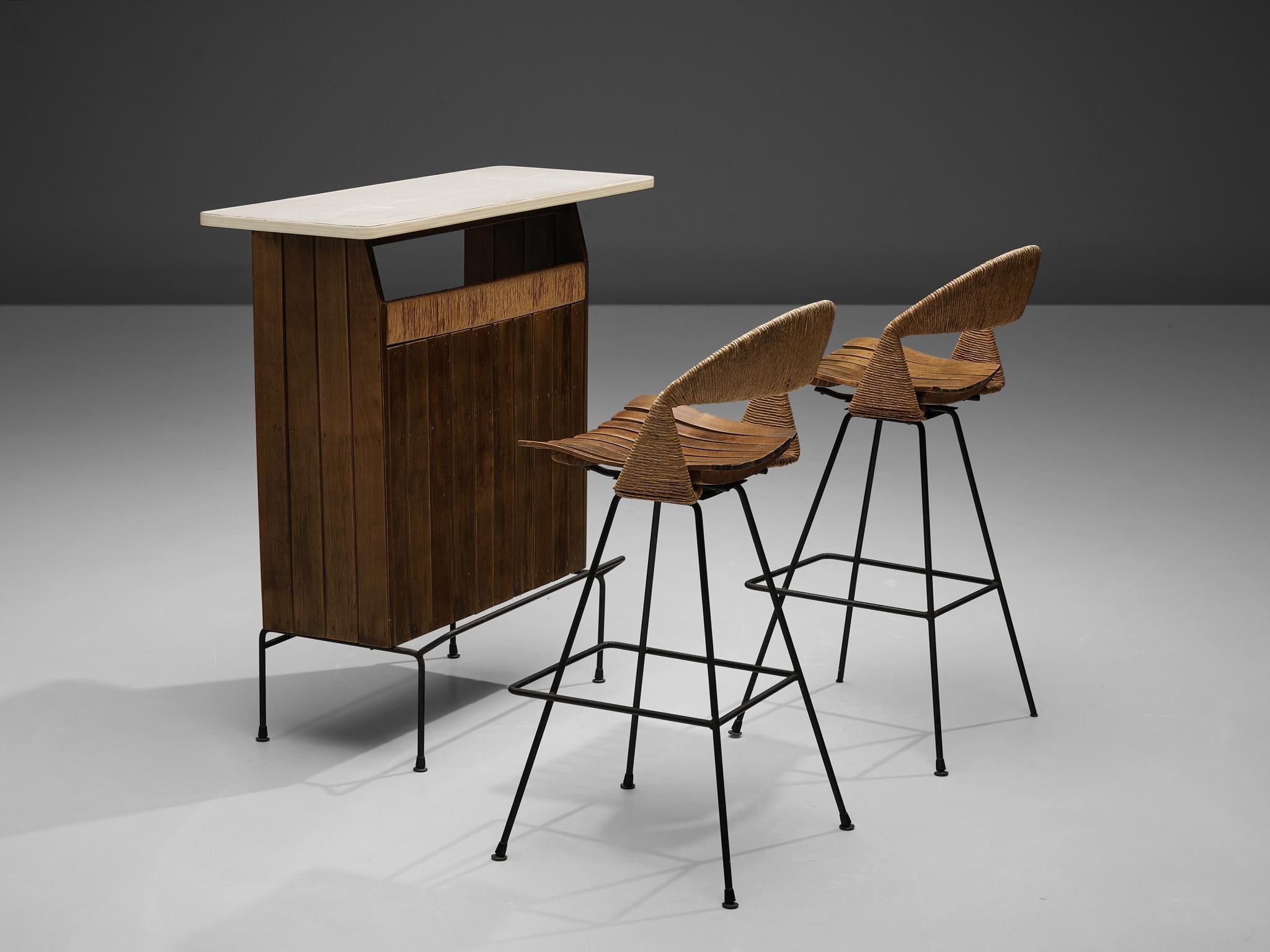 Arthur Umanoff for Raymor, dry bar, pair of bar stools, wood, iron, cane, United States, 1960s

The dry bar by Arthur Umanoff rests on a slim iron base that has a footrest in front. Vertical wooden slats end in a line highlighted in cane that flanks