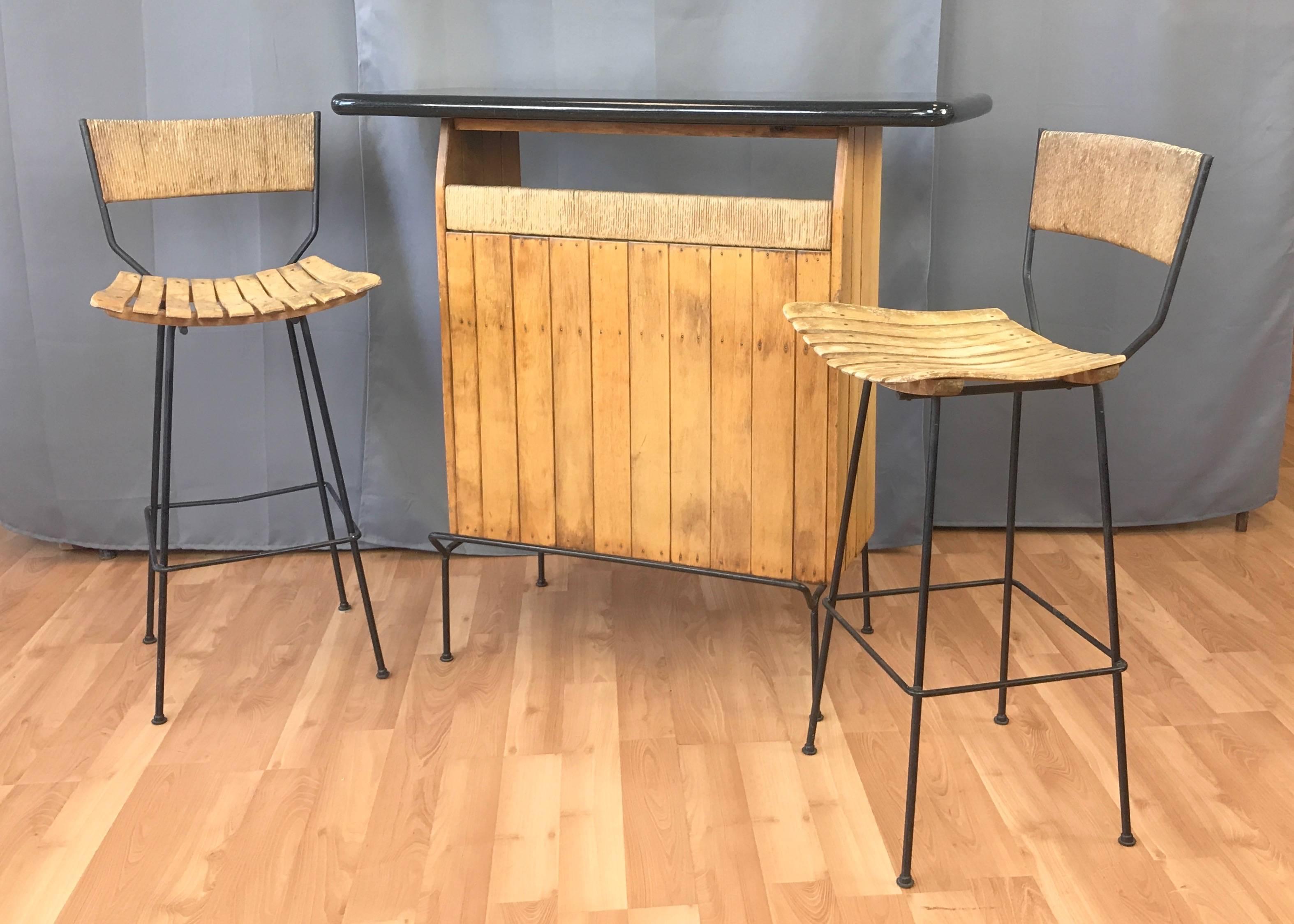A midcentury wrought iron, birch, and papercord dry bar with a pair of matching stools by Arthur Umanoff for Shaver Howard, and distributed by Raymor.

Bar has black wrought iron rod base with footrest, birch slat front and sides with