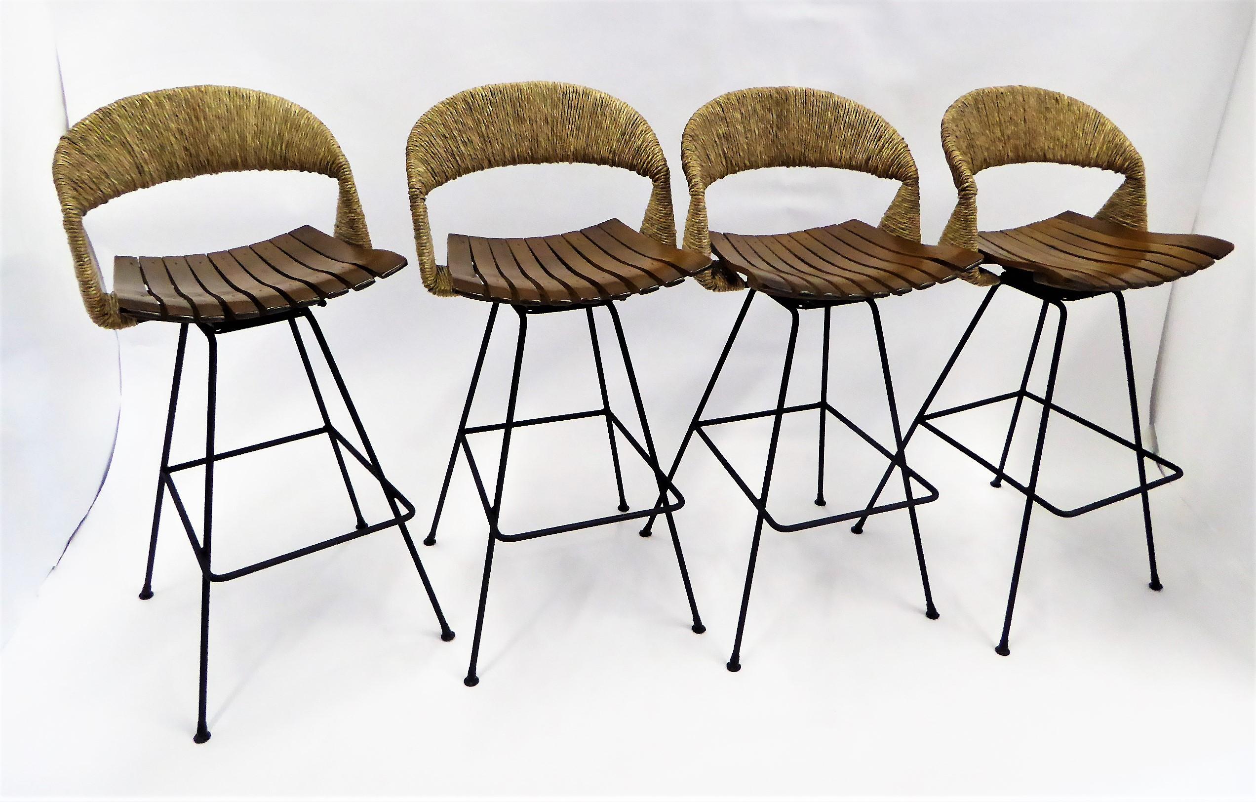 Designed by midcentury Master Arthur Umanoff and marketed by Raymor, here four Swiveling barstools featuring shapely handwoven raffia cord backs and wood slat seats all supported by a shaped Wrought Iron body with built in foot rests. New handwoven