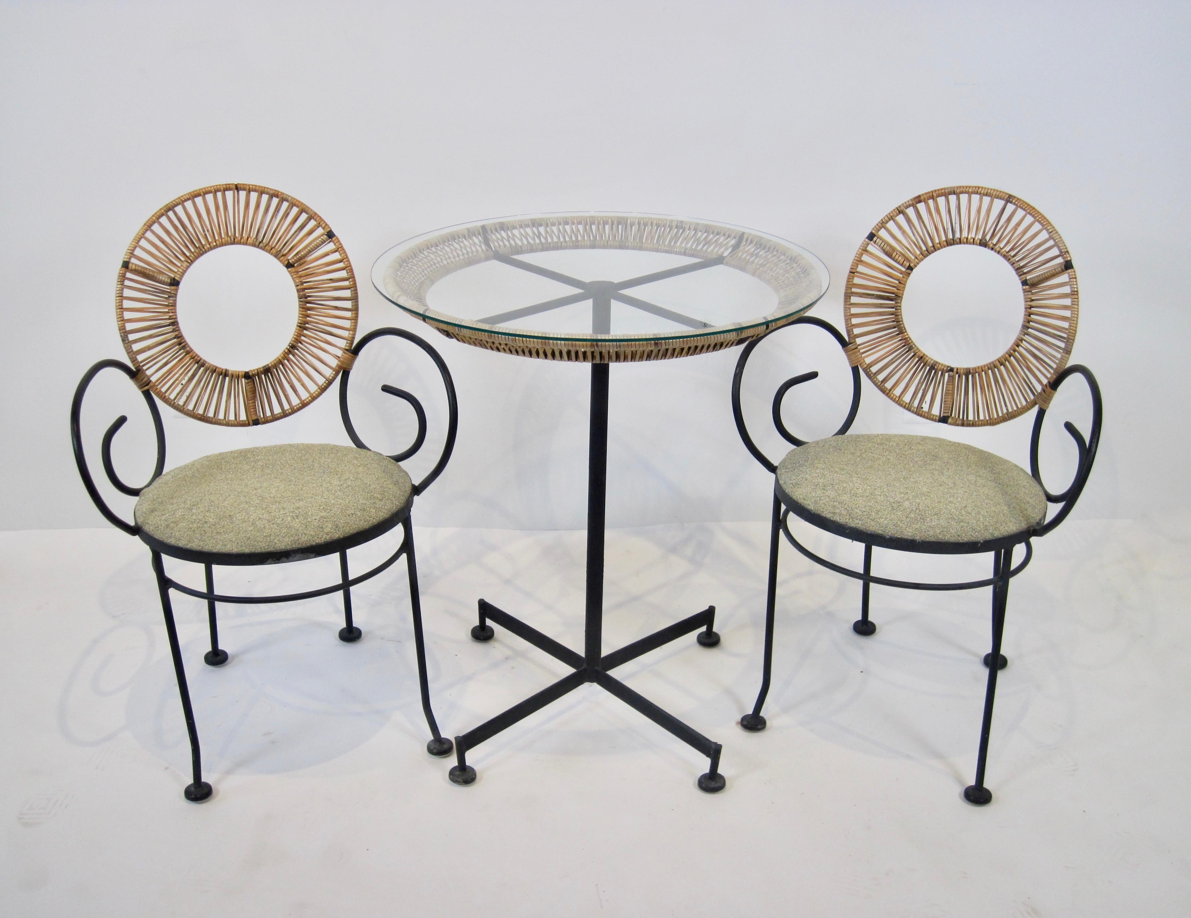A Mid Century wrought iron bistro set by Arthur Umanoff for Shaver Howard includes a pedestal table with four arm base and raffia wrap on table top with glass overlay. The two scroll arm chairs have raffia wrapped circular backs and round cushioned
